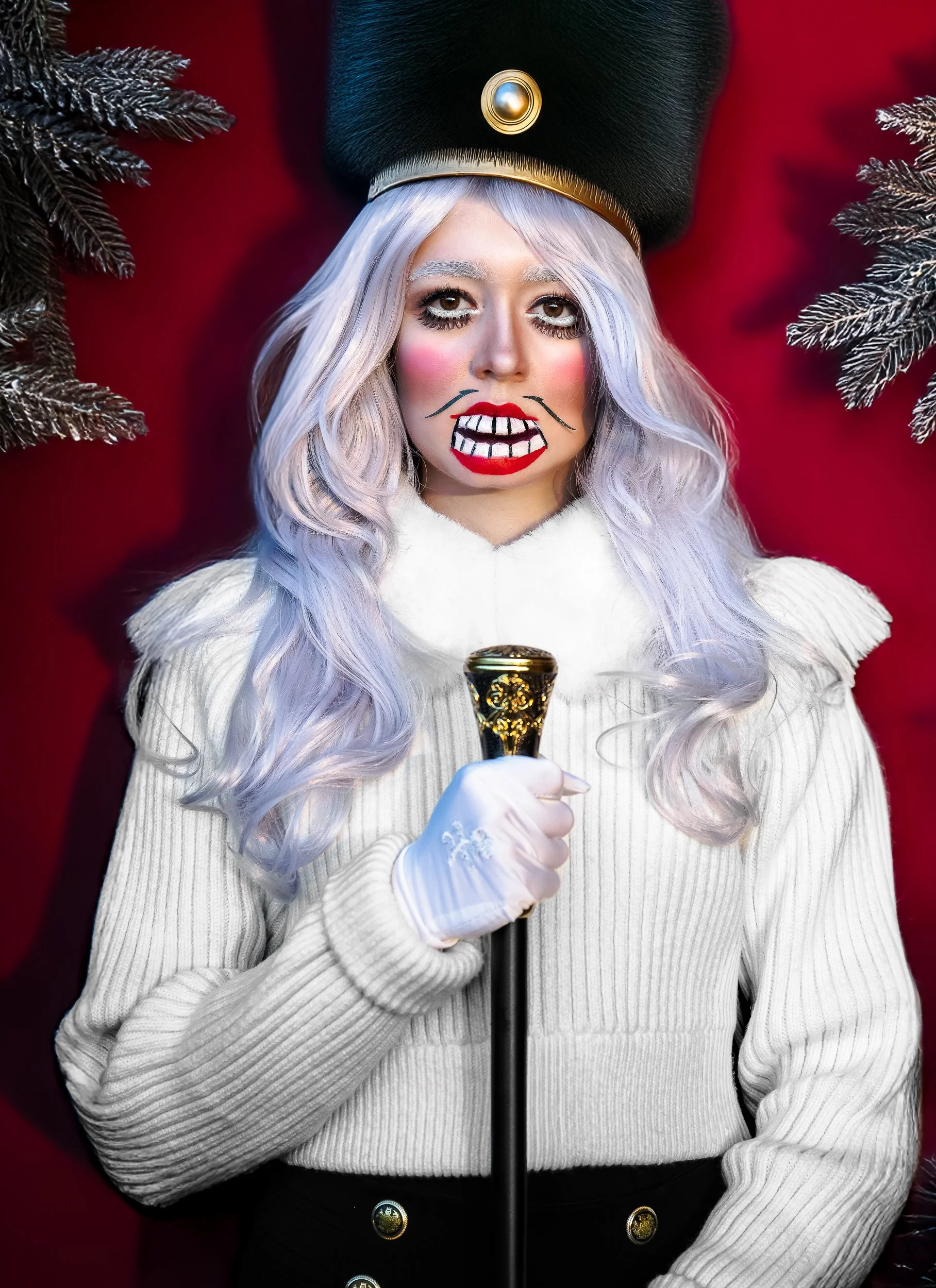 Une femme déguisée en personnage de Noël avec maquillage extravagant, portant un uniforme blanc, un chapeau noir orné d'une perle, tenant un bâton décoratif, avec un fond rouge et des branches de sapin argenté.