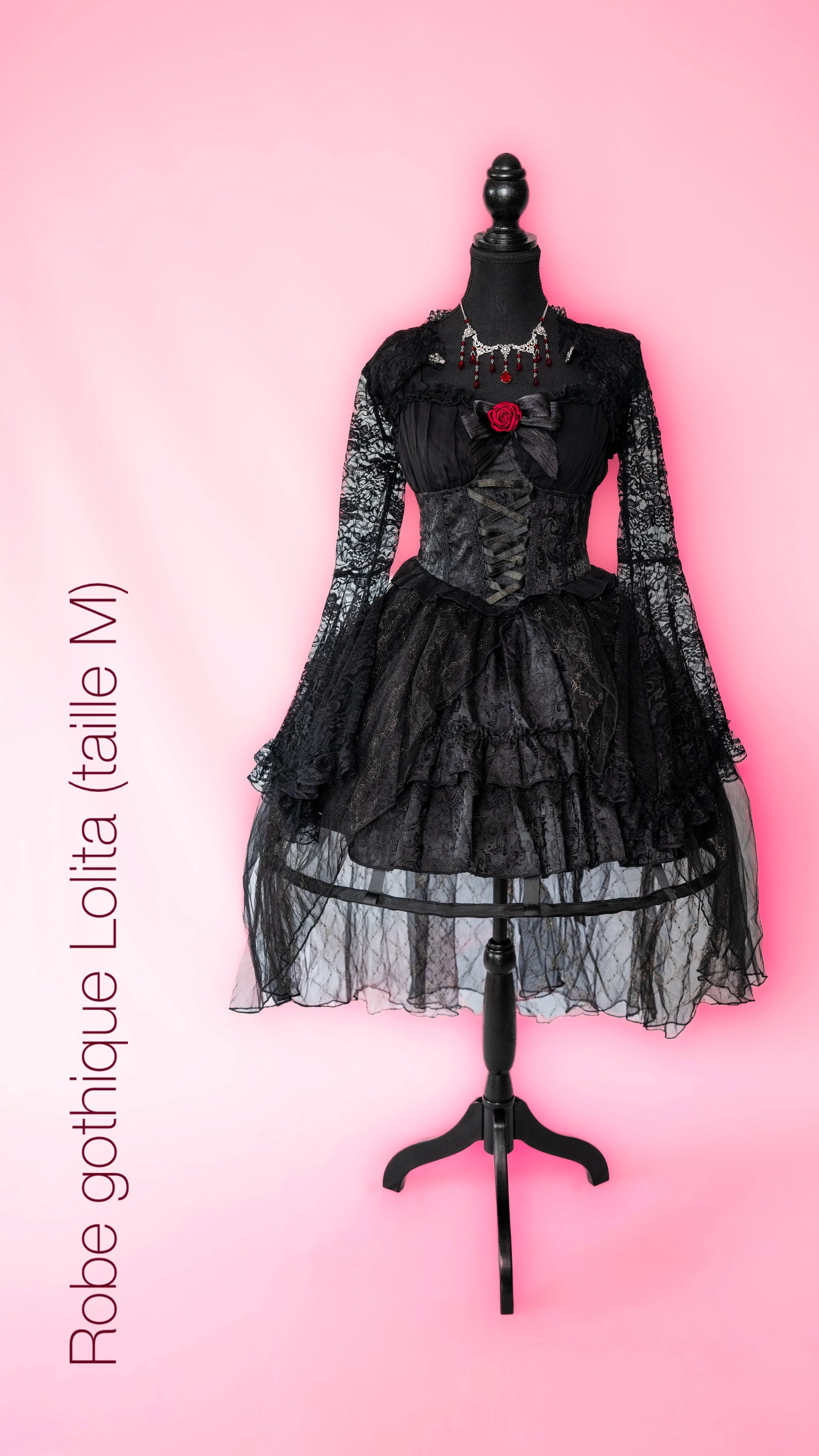 Robe gothique vintage noire avec dentelle, corset et détails roses, sur mannequin contre fond rose.