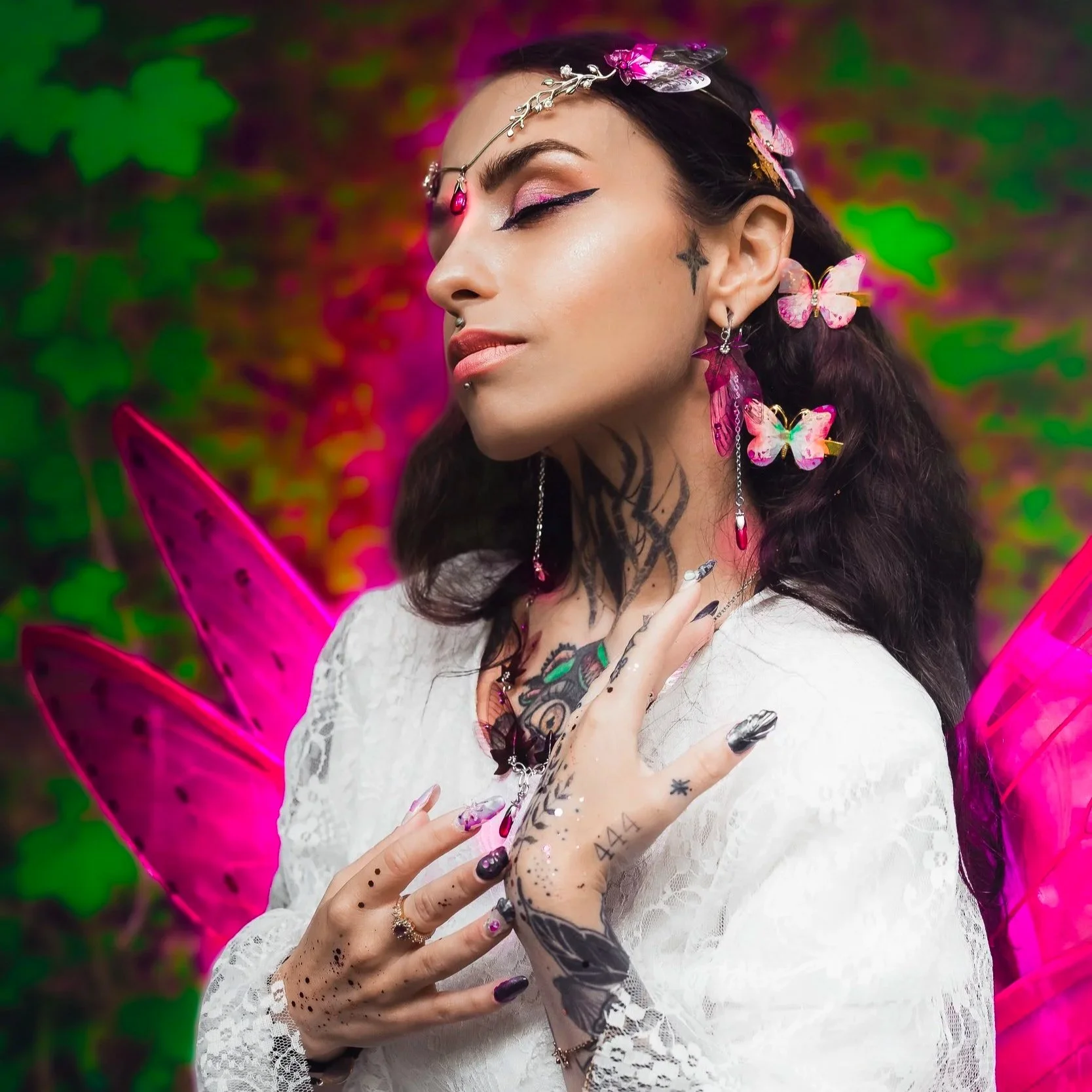 Une femme avec des ailes de fée roses, portant des accessoires de papillons et de bijoux, pose en fixant ses mains tatouées contre sa poitrine, avec un fond coloré de vert et de rose.