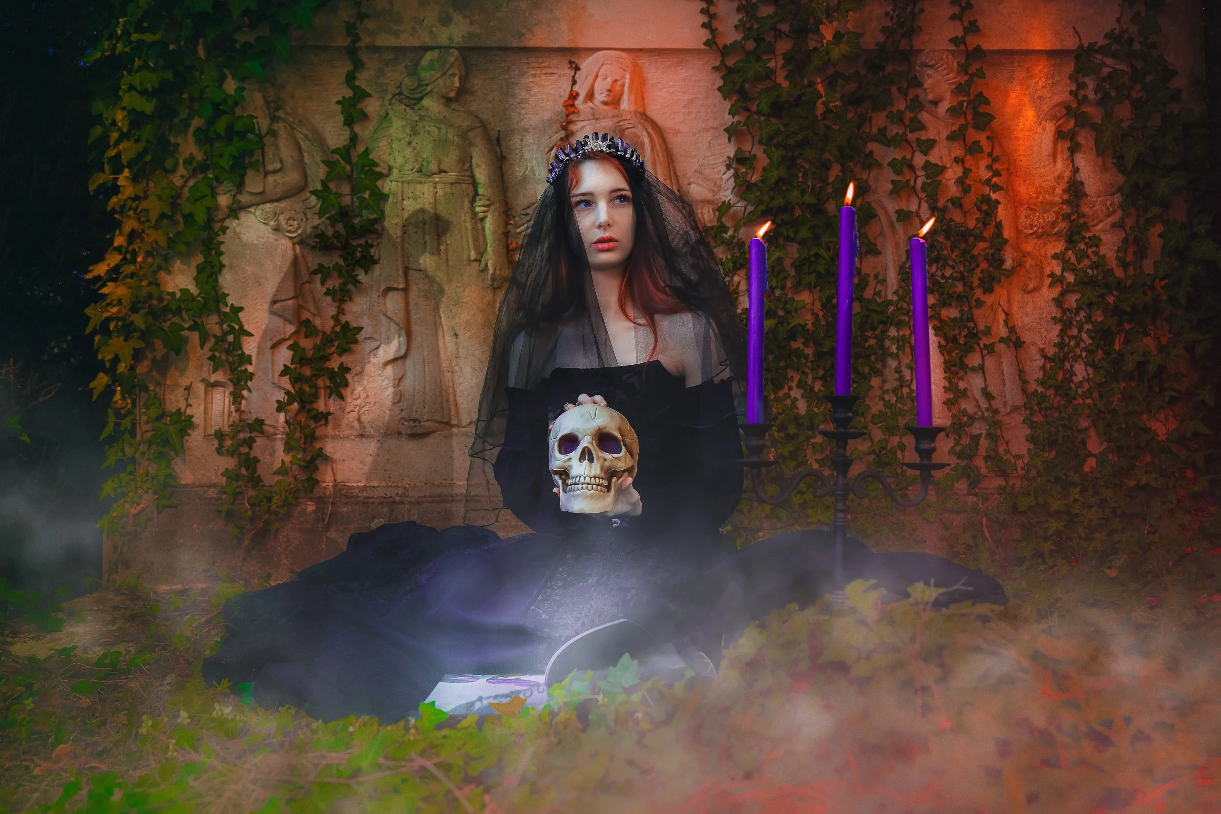 Une femme déguisée en sorcière ou en princesse sombre, tenant un crâne humain, assise devant un mur en pierre recouvert de vigne, avec des chandelles violettes allumées, dans une ambiance mystérieuse et effrayante, avec de la brume en premier plan.