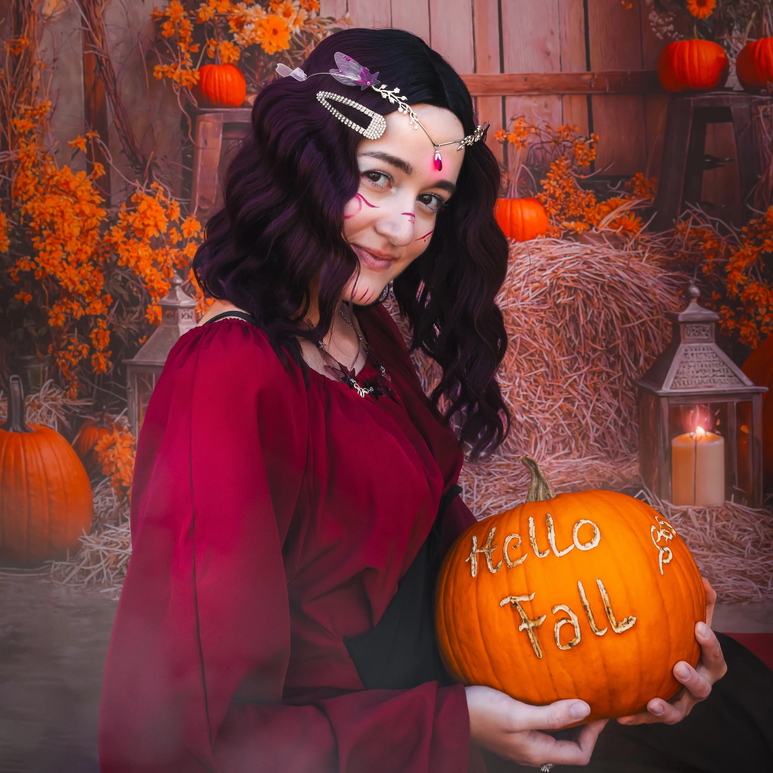 Une jeune femme habillée pour Halloween, tenant une citrouille avec l'inscription 'Hello Fall' en lettres blanches. Elle a des cheveux ondulés noirs avec des accessoires, et un maquillage effrayant. En arrière-plan, décoration automnale avec des citrouilles, des lanternes et des fleurs orange.