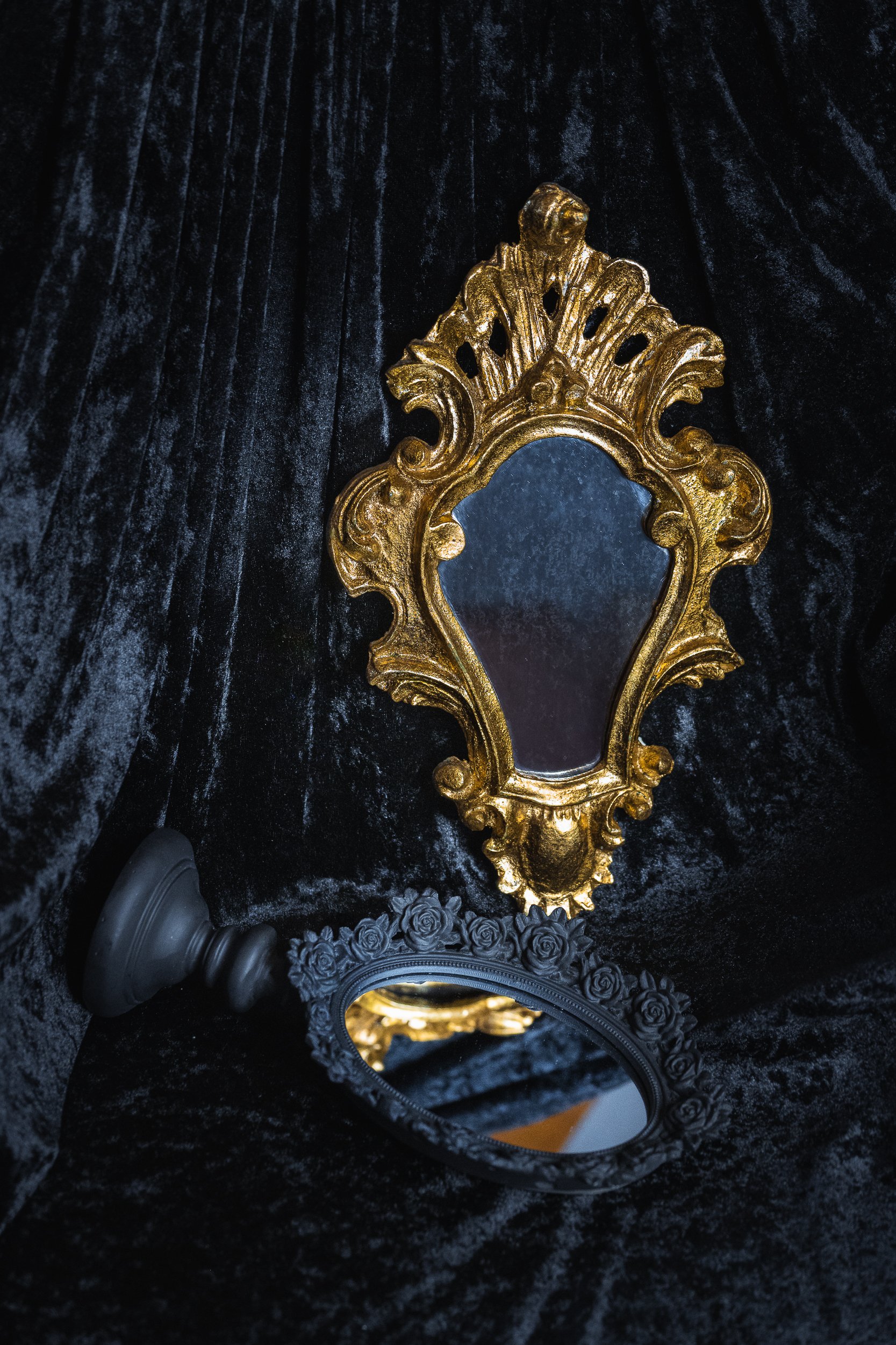 Un miroir doré complexe posé devant un miroir noir à décor de roses, tous deux sur un fond de tissu noir velvet.