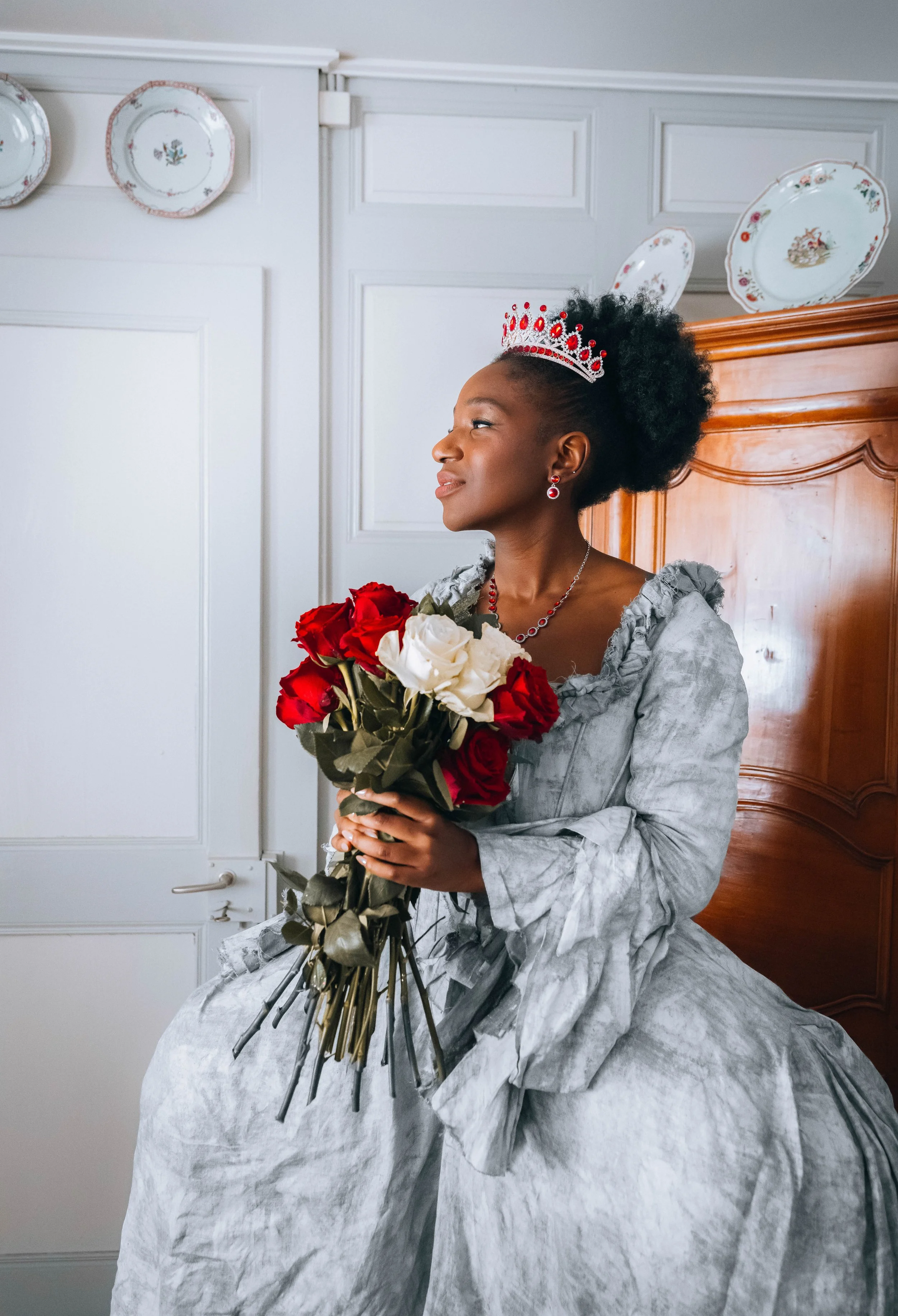 Une femme noire en robe d'époque, portant une couronne et des bijoux, tient un bouquet de roses rouges et blanches, assise dans une pièce avec moquette en bois et assiettes décoratives accrochées au mur.