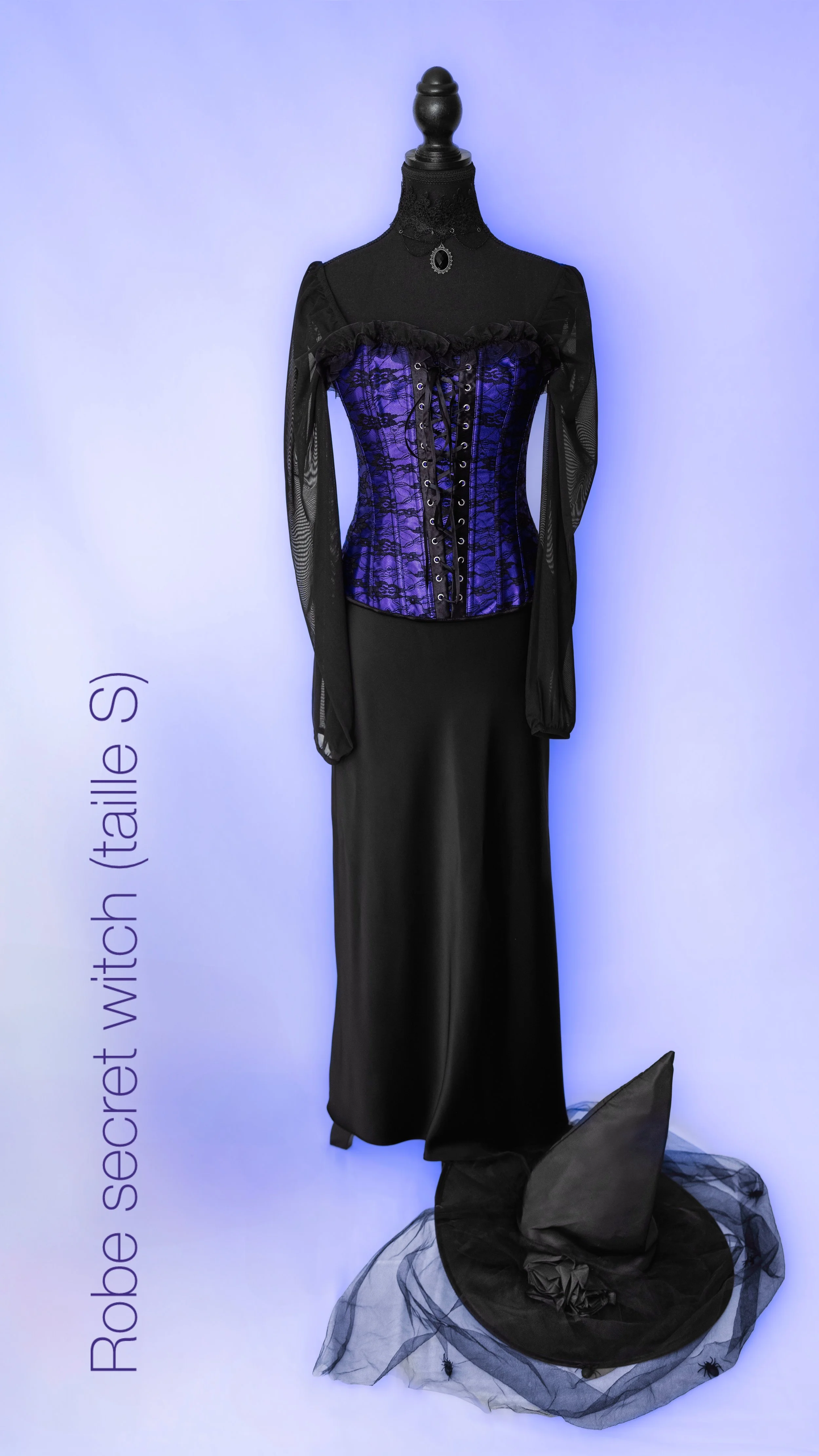 Une robe de sorcière secrète avec un corset violet et une cape noire, exposée sur un mannequin, accompagnée d'un chapeau de sorcière noir sur un plateau décoratif contenant du tulle noir et des araignées noires.