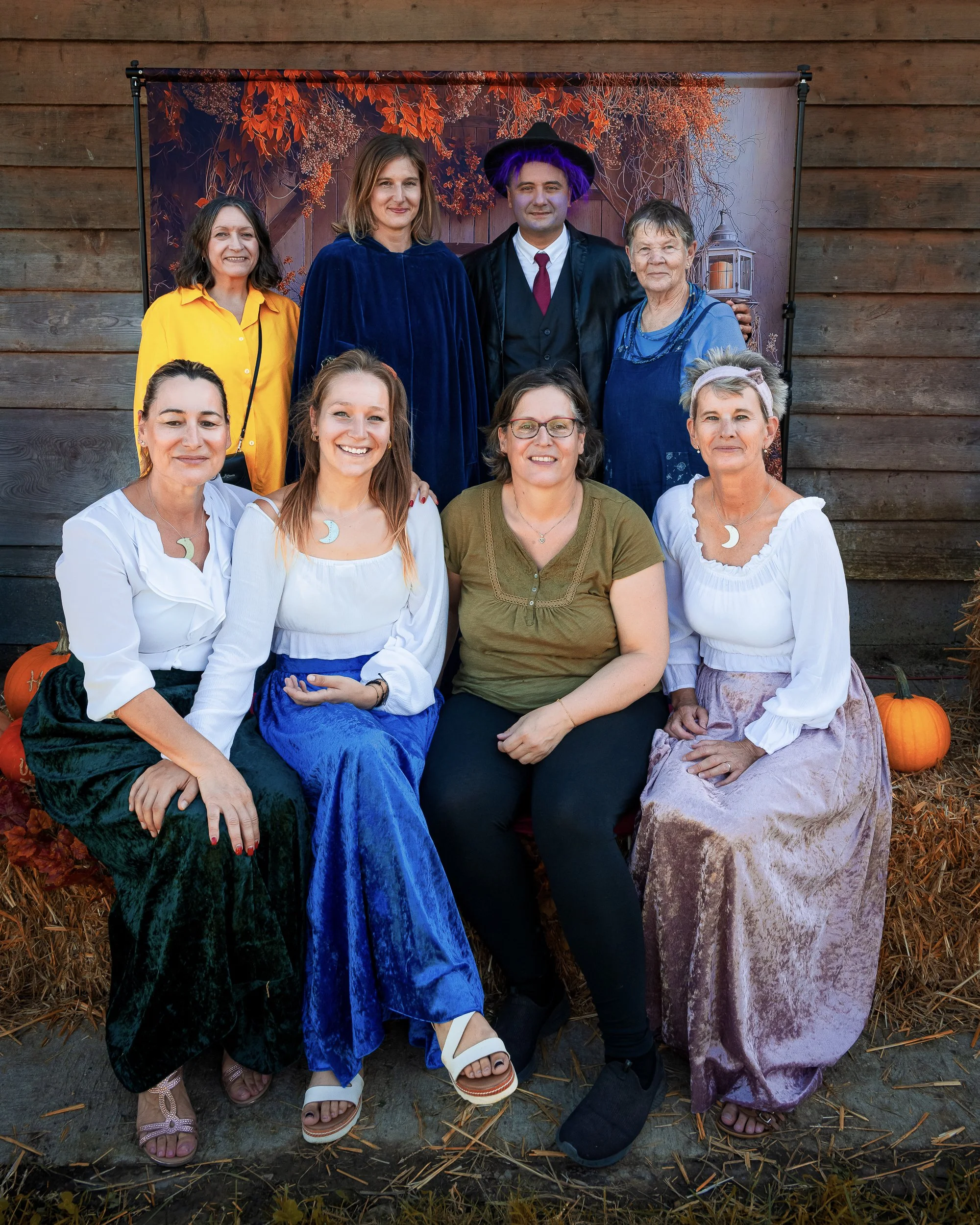 Groupe de neuf personnes posant pour une photo à l'extérieur, devant un fond décoratif avec des feuilles d'automne. Certaines portent un déguisement d'Halloween, avec un sorcier, tandis que d'autres portent des vêtements décontractés ou vintages. Il y a des citrouilles à leurs côtés et un sol recouvert de paille.