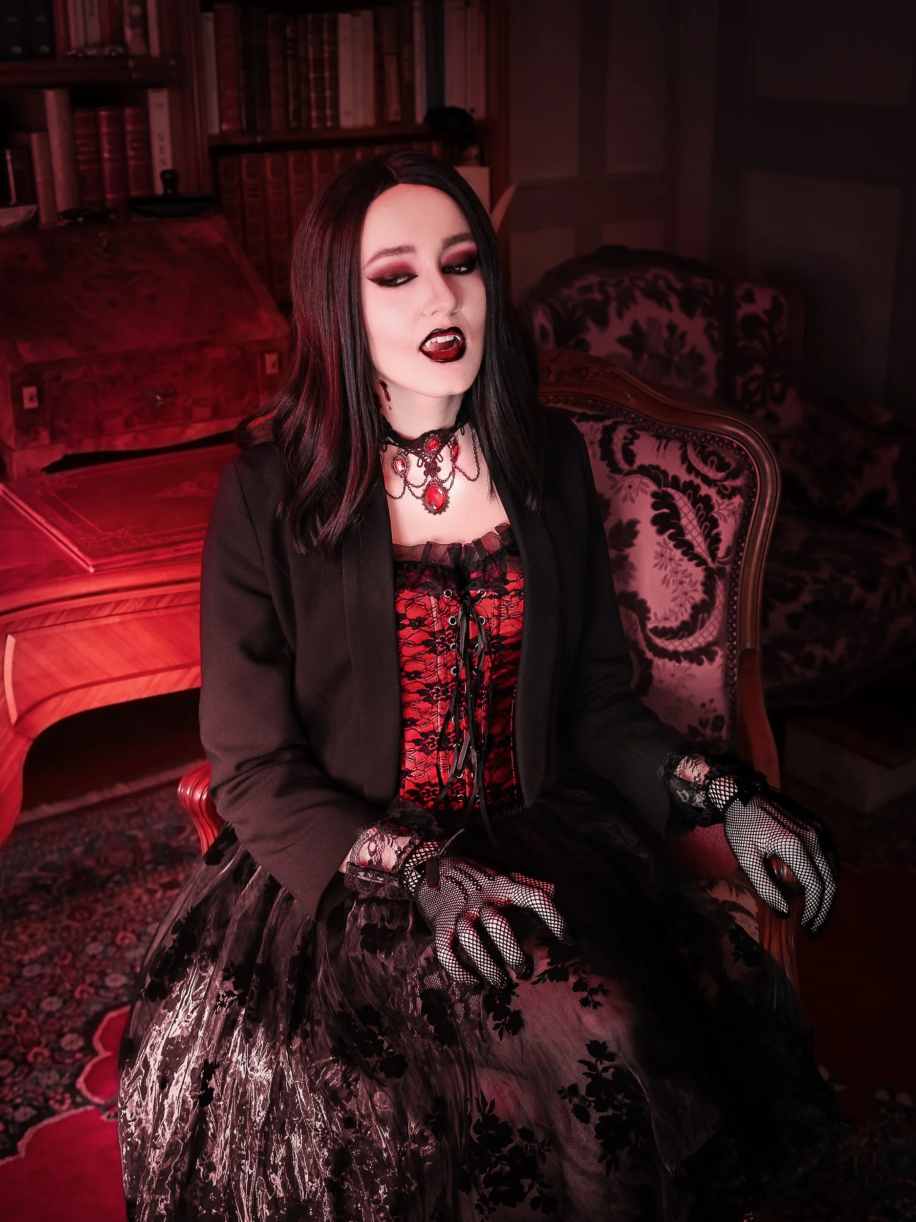 Une femme vêtue d'une tenue gothique noire et rouge, portant des gants en filet, assise dans un salon avec mobiliers anciens et étagère pleine de livres.