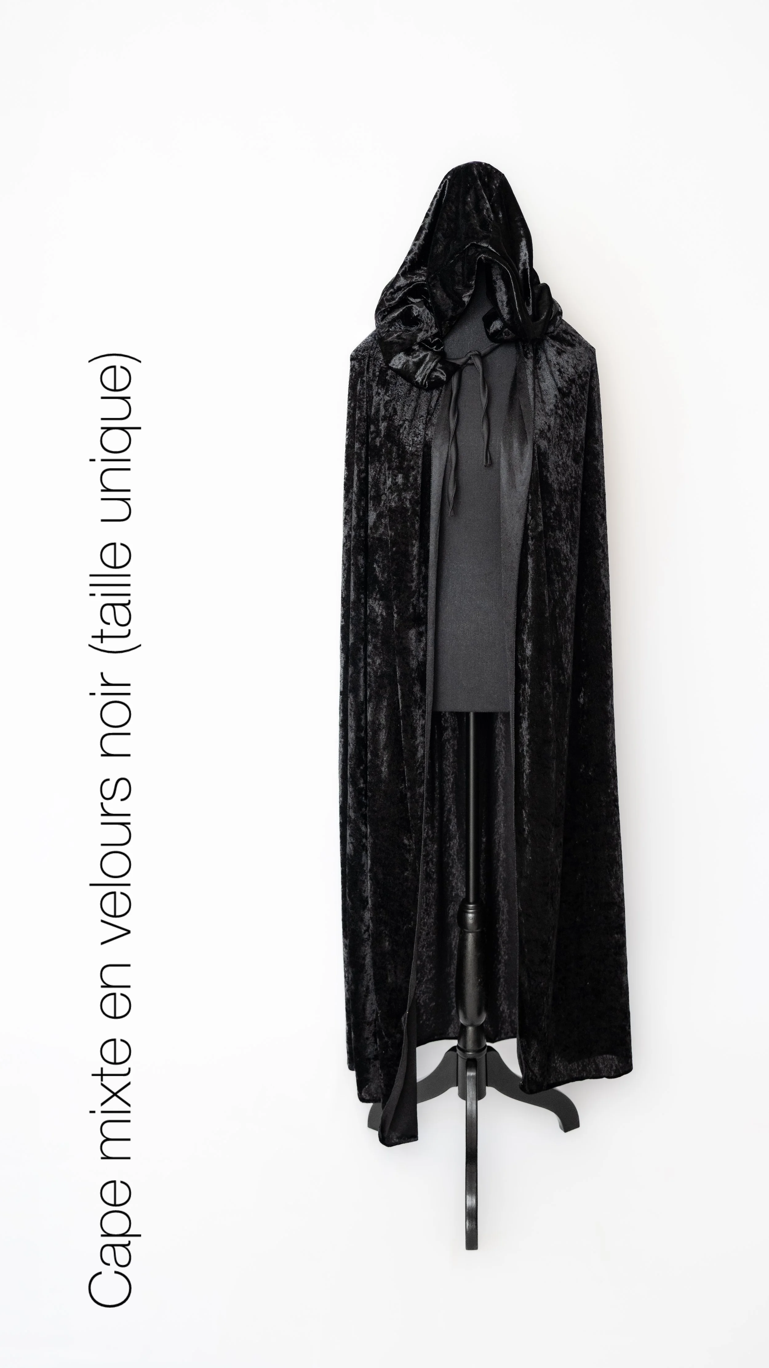 Mannequin habillé d'une cape noire en velours, longue et avec un capuchon, contre un mur blanc.