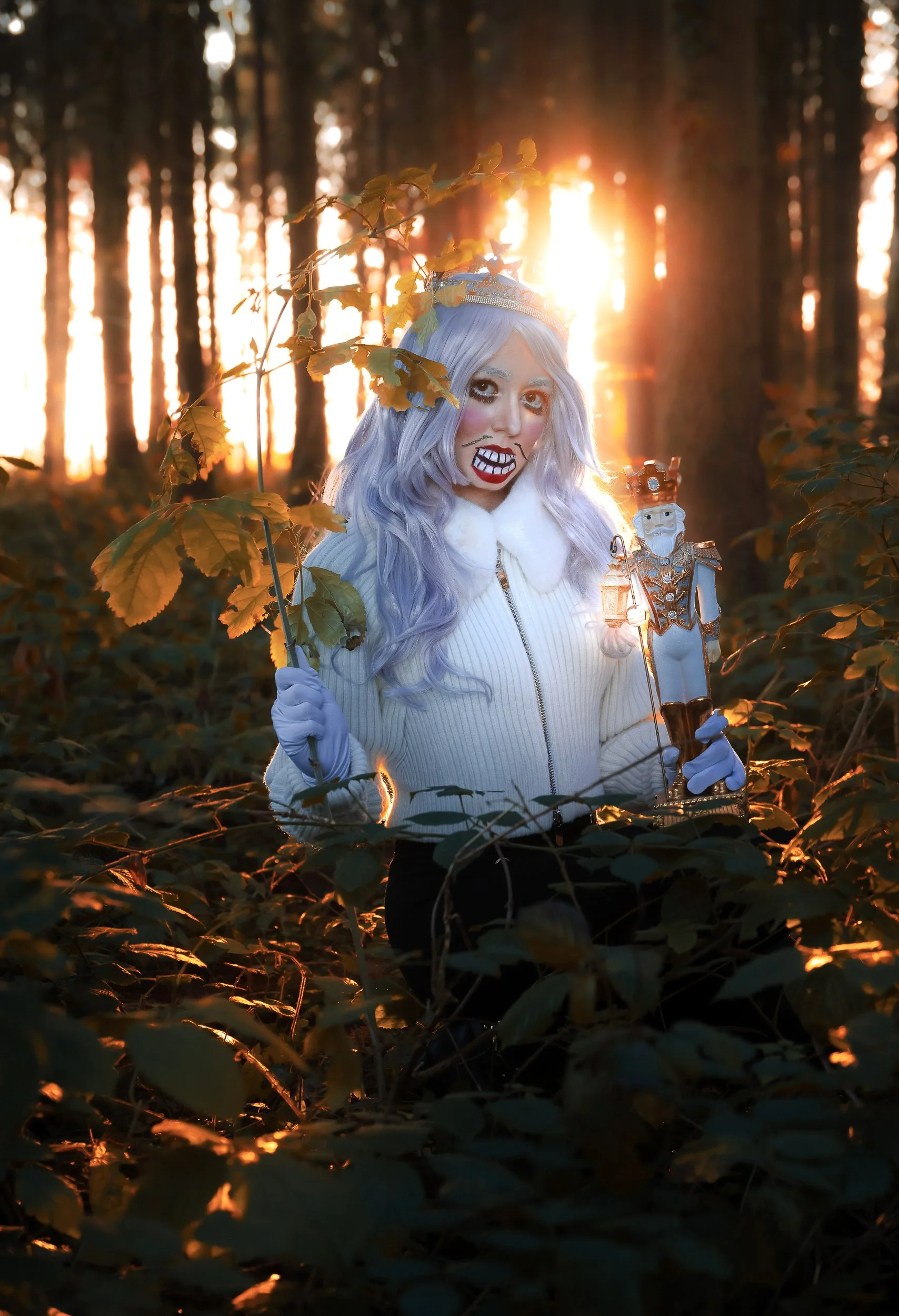 Une femme déguisée en zombie dans une forêt au coucher du soleil, tenant une poupée de noël et une branche. Elle a des cheveux violets, un visage maquillé en zombie et porte un pull blanc avec une fermeture éclair.