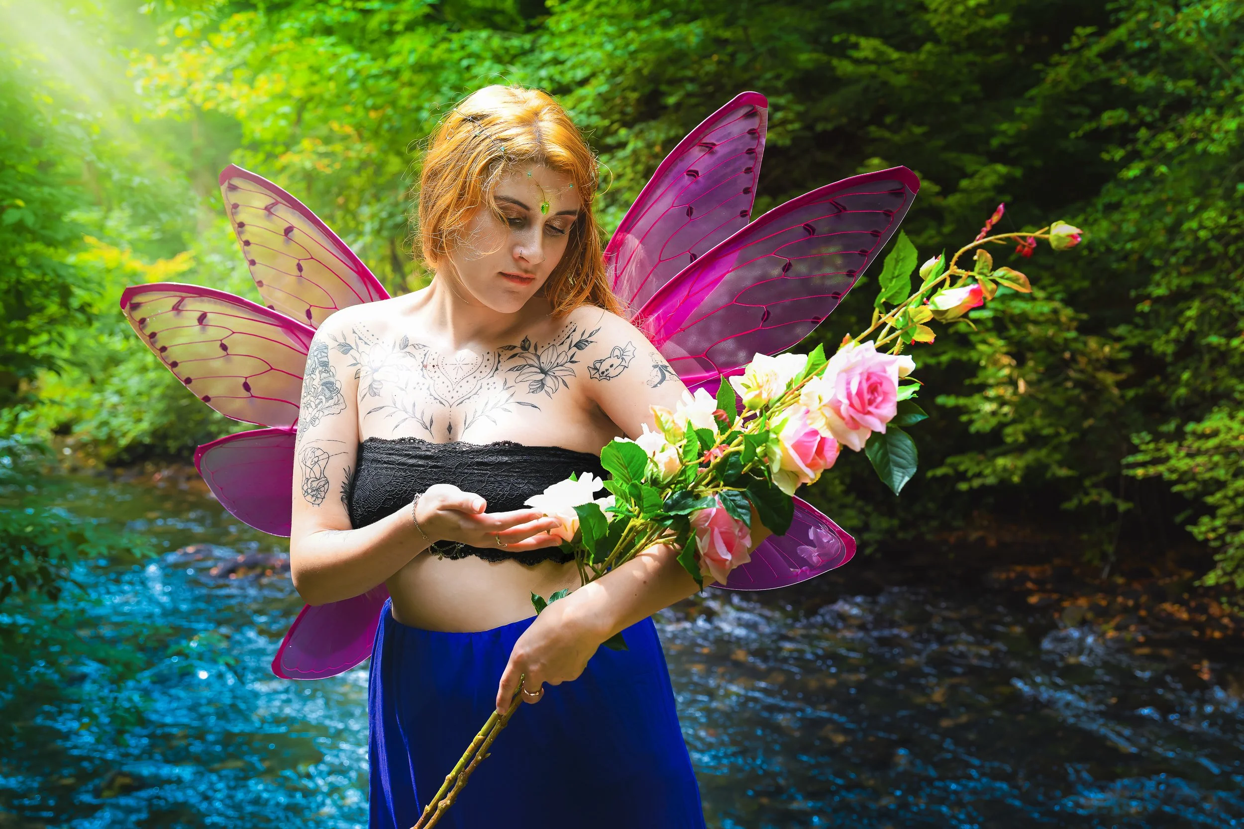 Une femme avec des ailes colorées de fée, portant un bustier noir, des tatouages visibles sur les bras et la poitrine, tient une branche de roses dans un environnement verdoyant près d'une rivière.