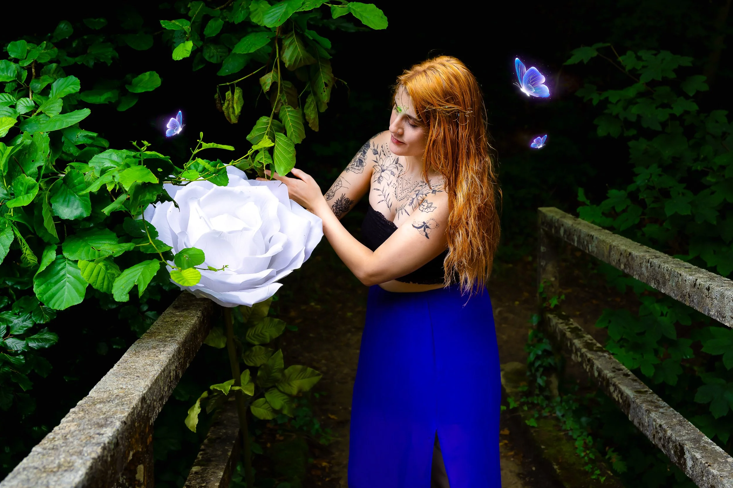 Une femme aux cheveux roux, tatouages visibles, portant une robe noire et une jupe bleue, se tient sur un sentier entouré de végétation, près d'un arbre avec une structure en papier blanc représentant une fleur, et regarde attentivement la plante.