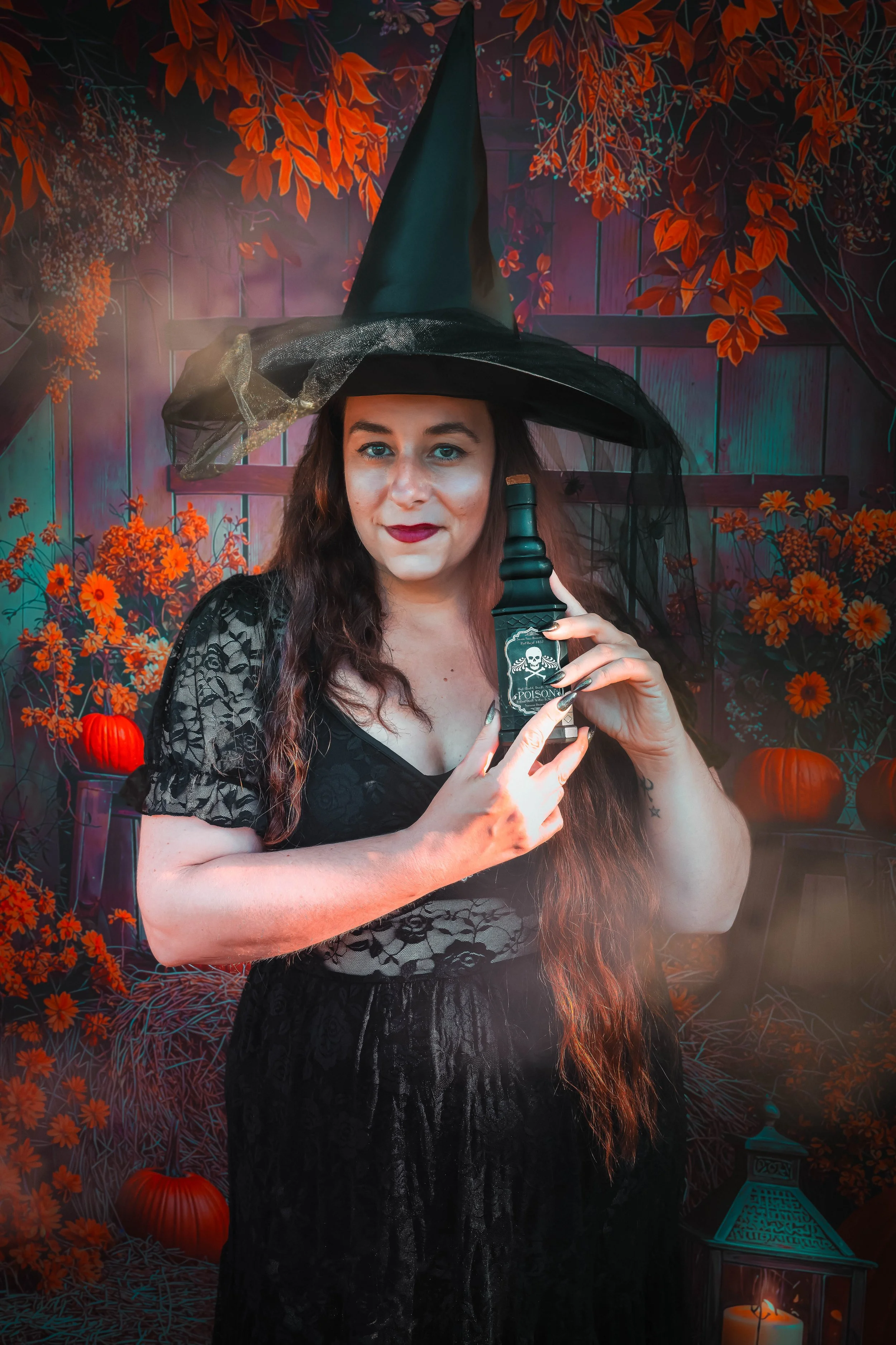 Une femme déguisée en sorcière avec un chapeau pointu noir, tenant une bouteille avec un symbole de danger, se tient devant un décor Halloween avec des citrouilles orange, des fleurs d'automne et des lanternes.