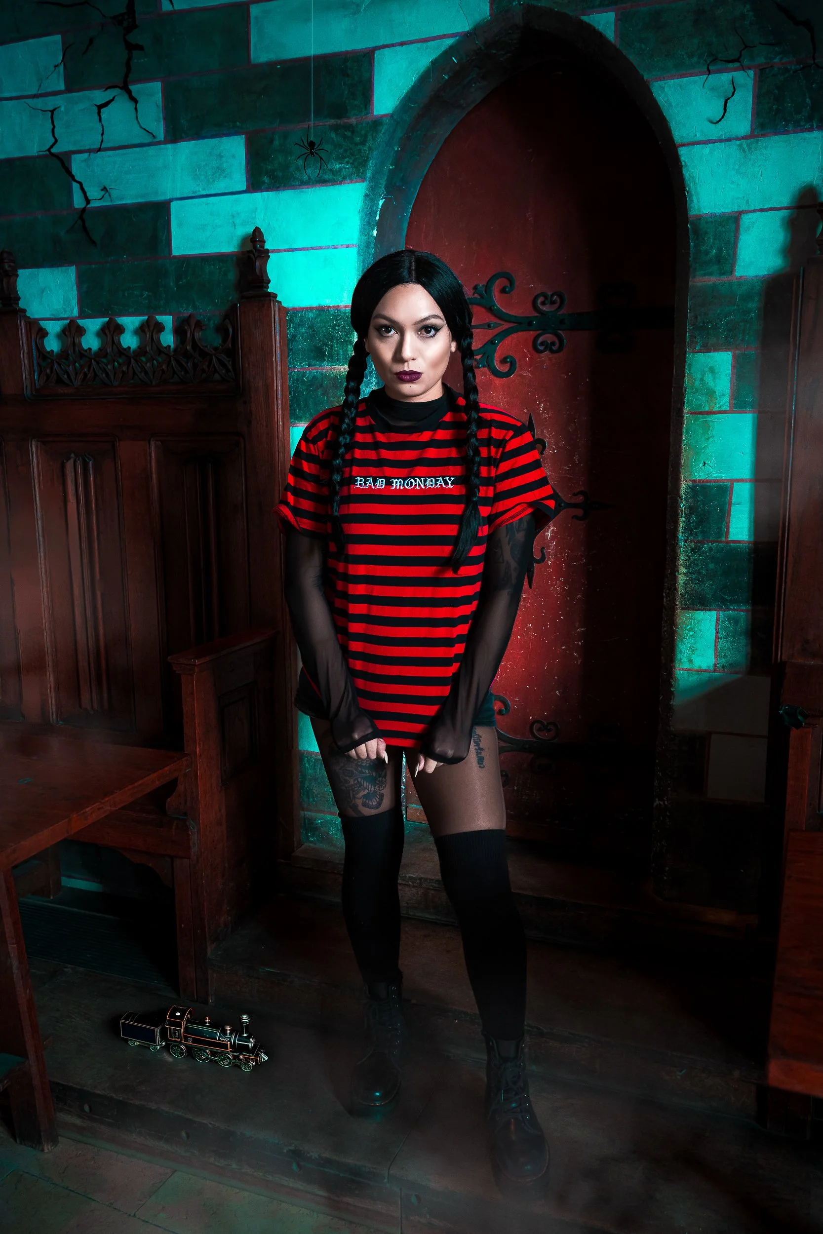 Femme avec des cheveux noirs tressés, portant un t-shirt rayé rouge et noir, des collants noirs avec motif floral, des bottes noires, dans une pièce sombre avec mur en pierre, une petite locomotive sur le sol, et décor d'araignées au plafond.