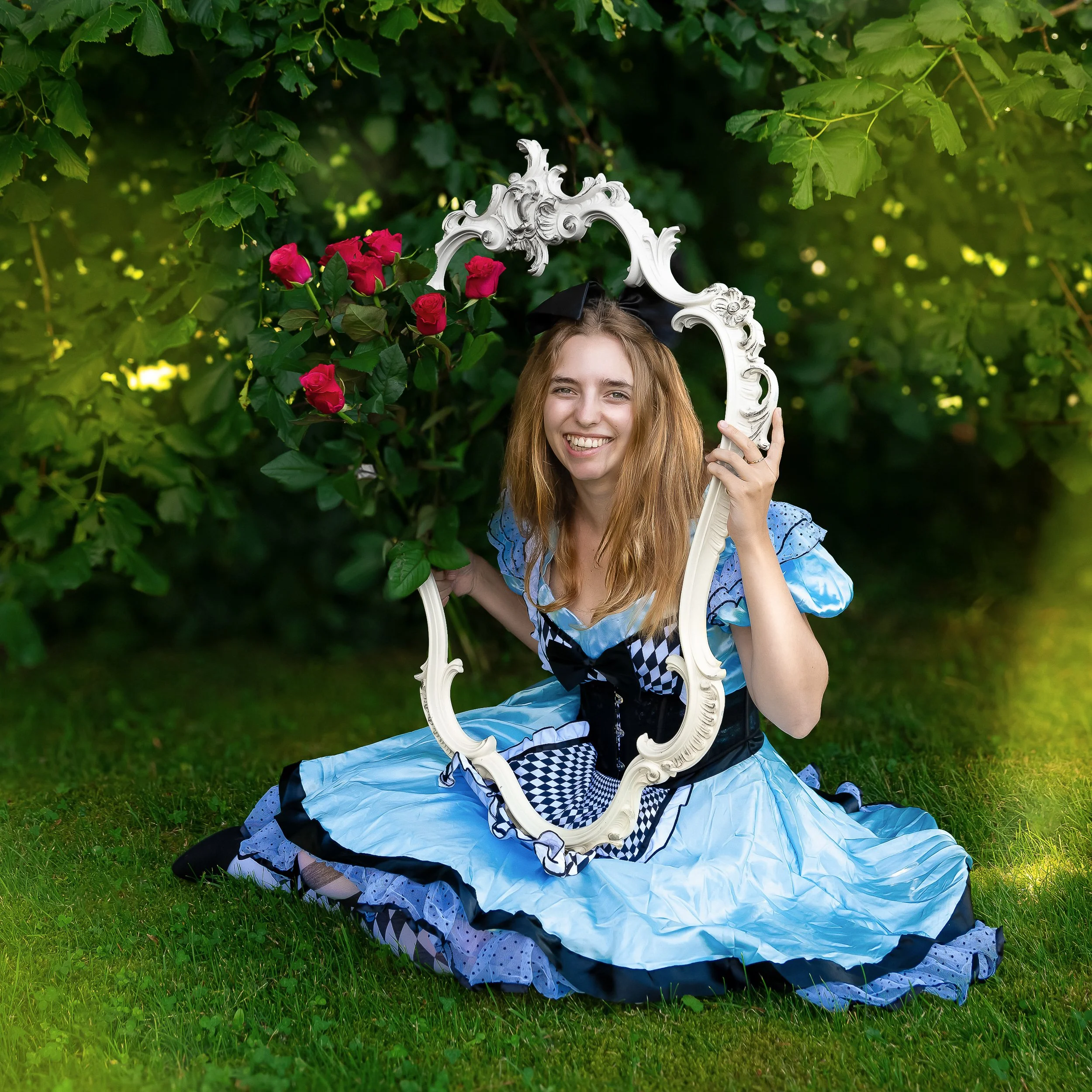 Jeune femme souriante en costume de conte de fées, assise sur l'herbe, tenant un grand miroir blanc orné, entourée de feuilles vertes et de fleurs roses.