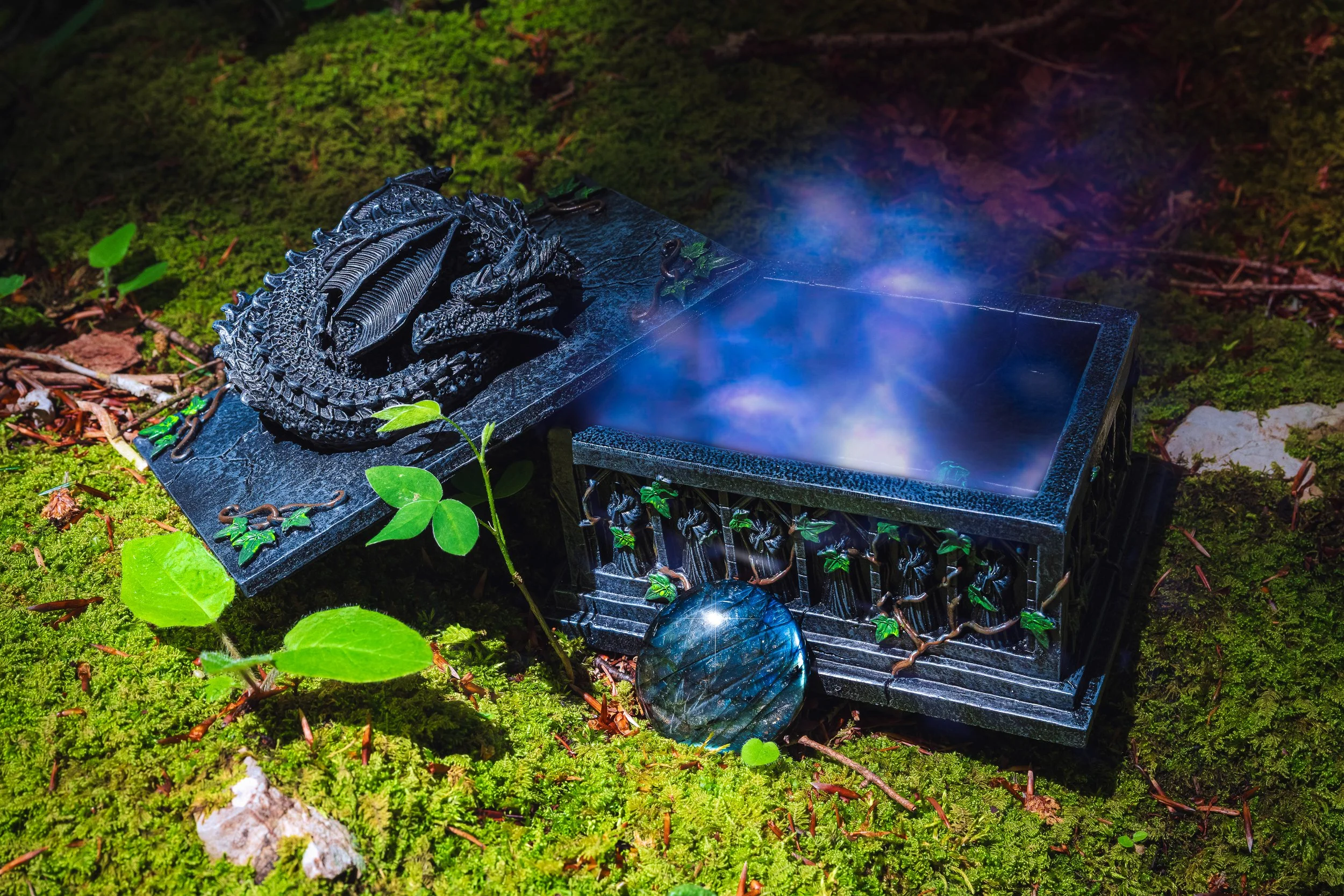 Coffre en forme de dragon noir avec fumée bleue sortant, posé sur une surface de mousse verte avec des feuilles.