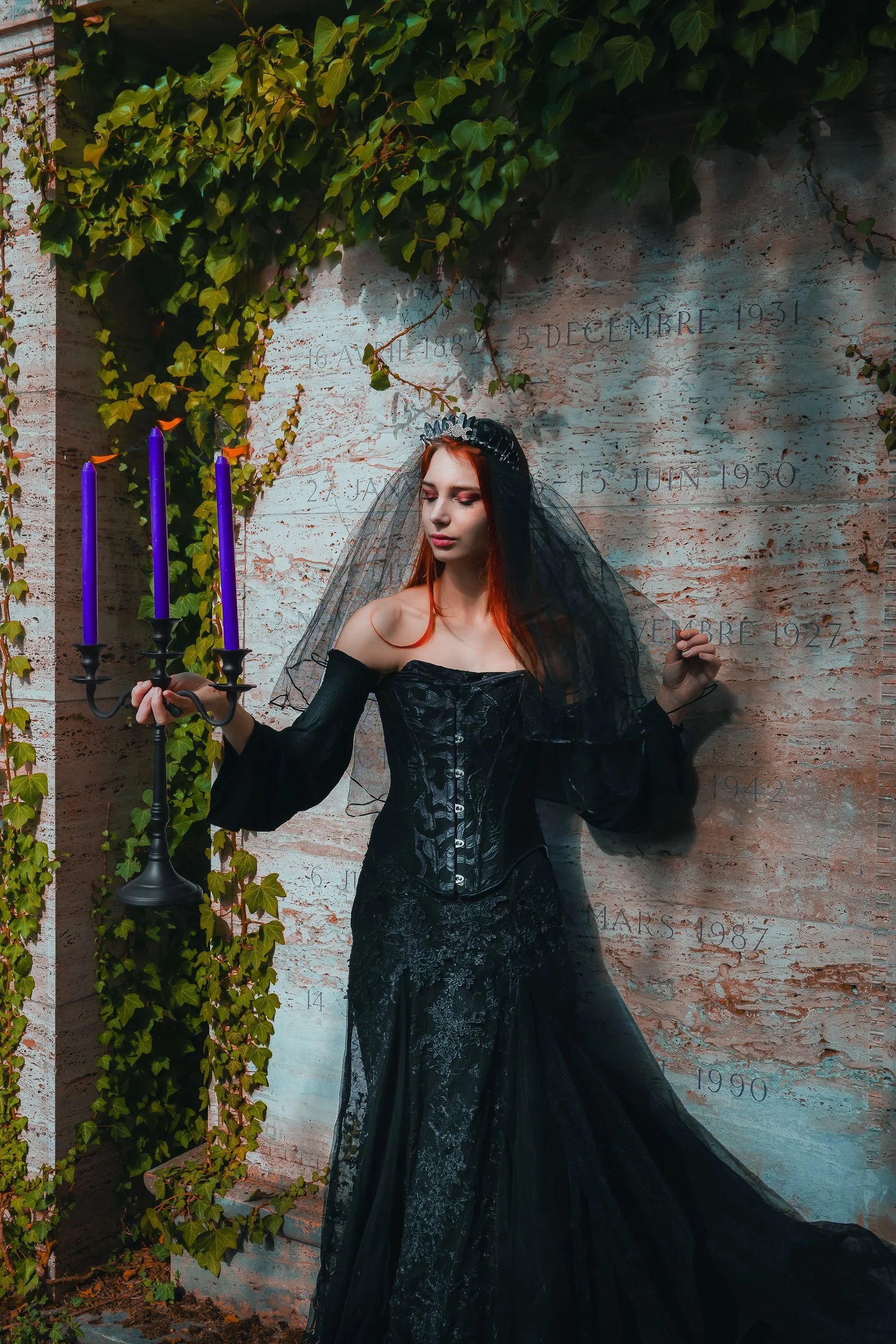 Jeune femme en robe noire élégante avec un voile noir et une couronne, tenant un chandelier avec des bougies violettes, posant devant un mur en pierre avec des inscriptions de dates.