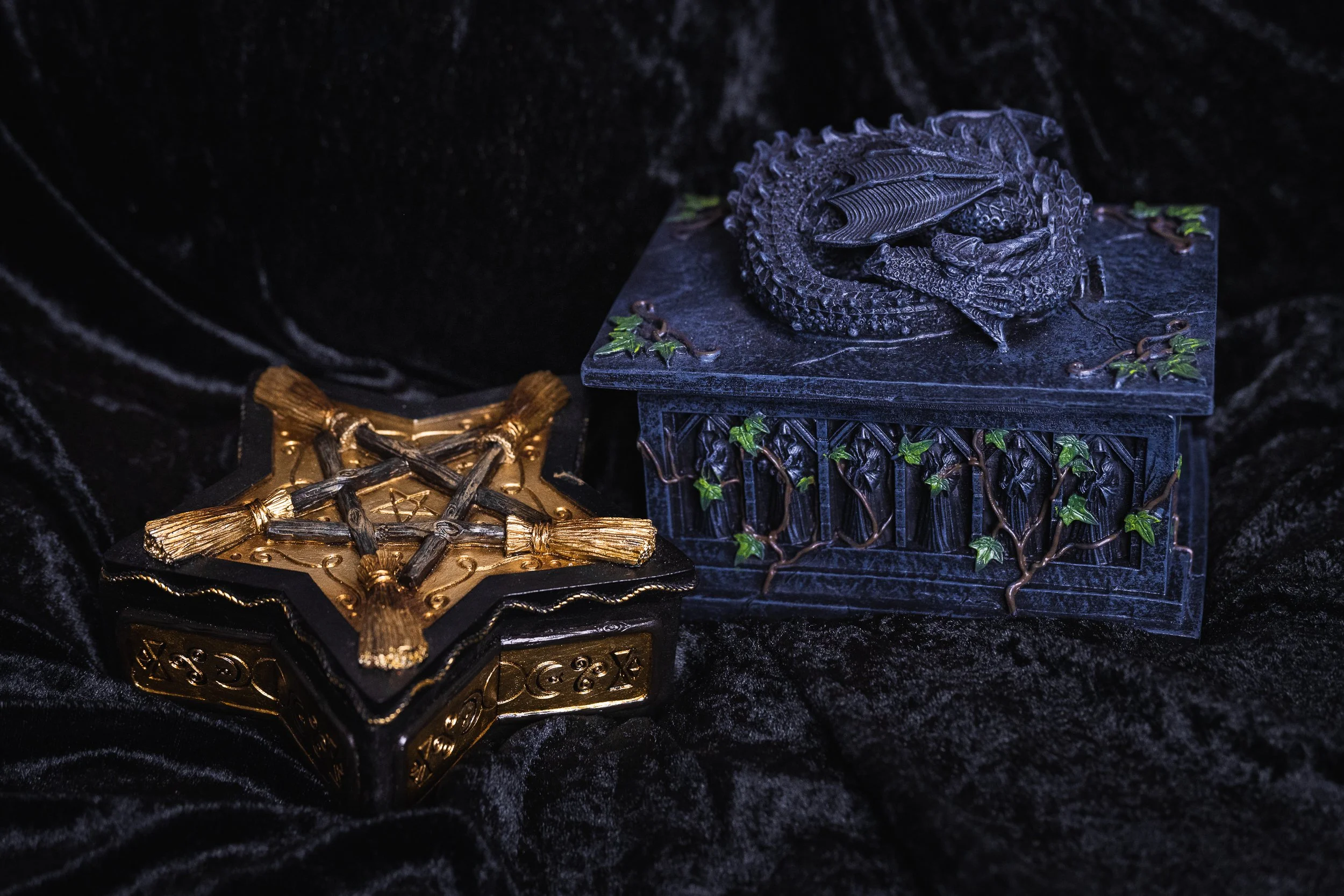 Deux coffrets décoratifs, un en or avec une croix, et l'autre en métal noir avec un dragon, posés sur un tissu noir velours.