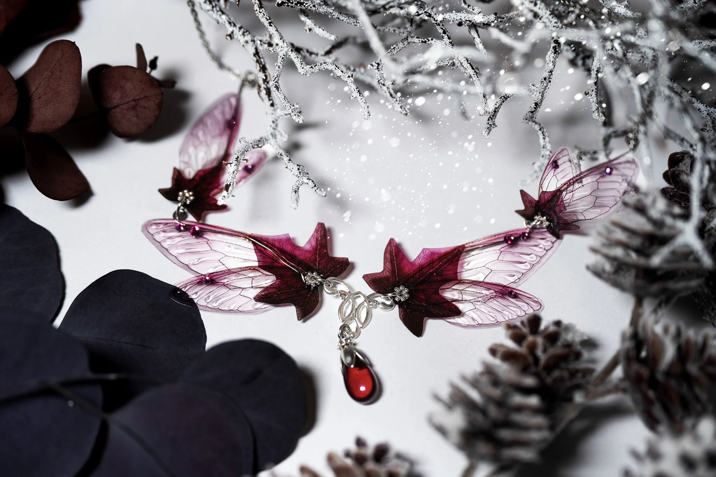 Collier en forme de papillons et feuilles en verre rose et rouge, avec une pierre rouge suspendue, entouré de branches blanches givrées, brindilles et feuilles sèches sur fond blanc.
