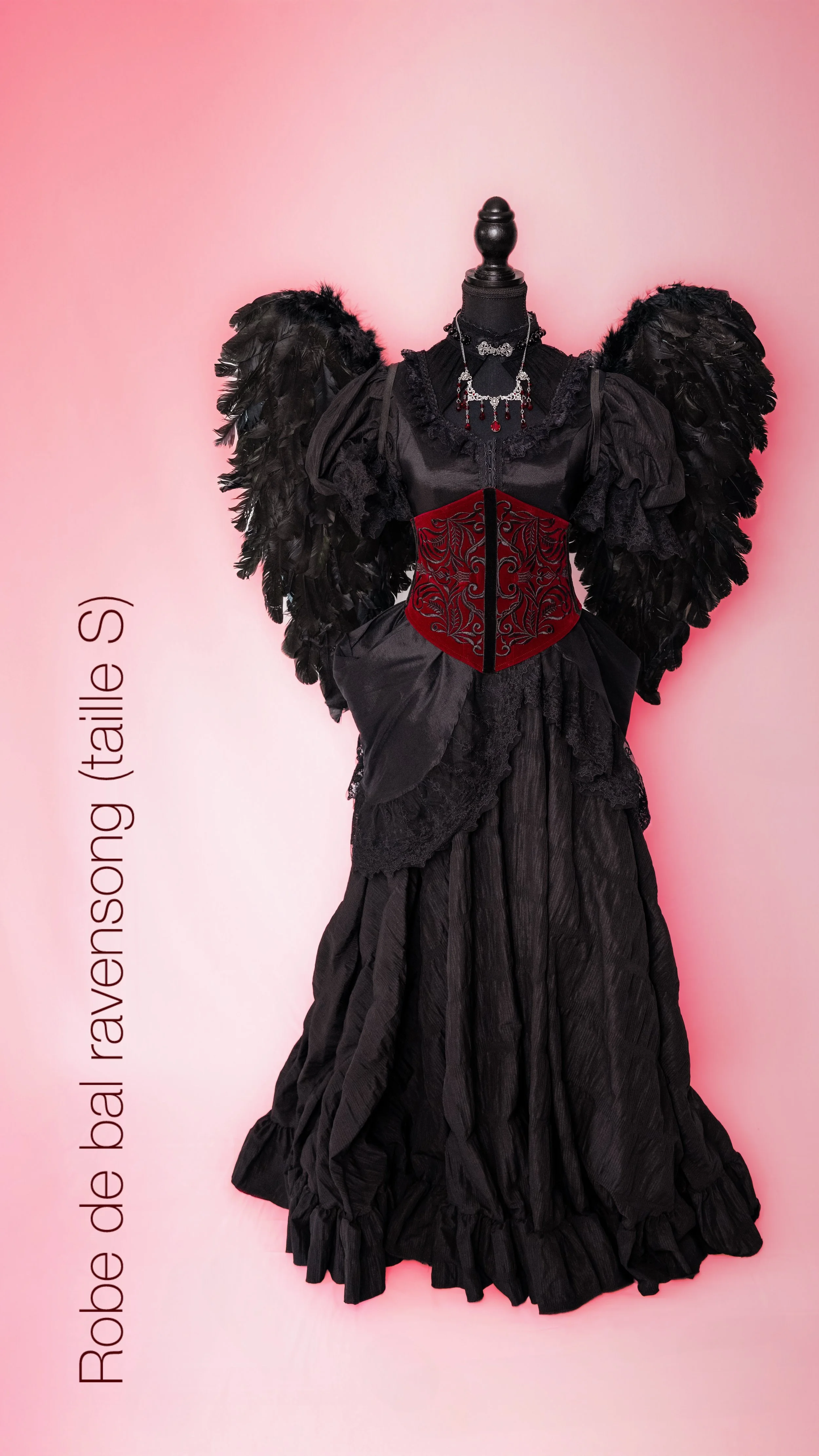 Une robe de bal ravensong (Italie) noire avec des détails en dentelle, un corset rouge brodé, des manches volumineuses en tissu noir, un col en dentelle, une chaîne de bijoux, sur fond rose dégradé.