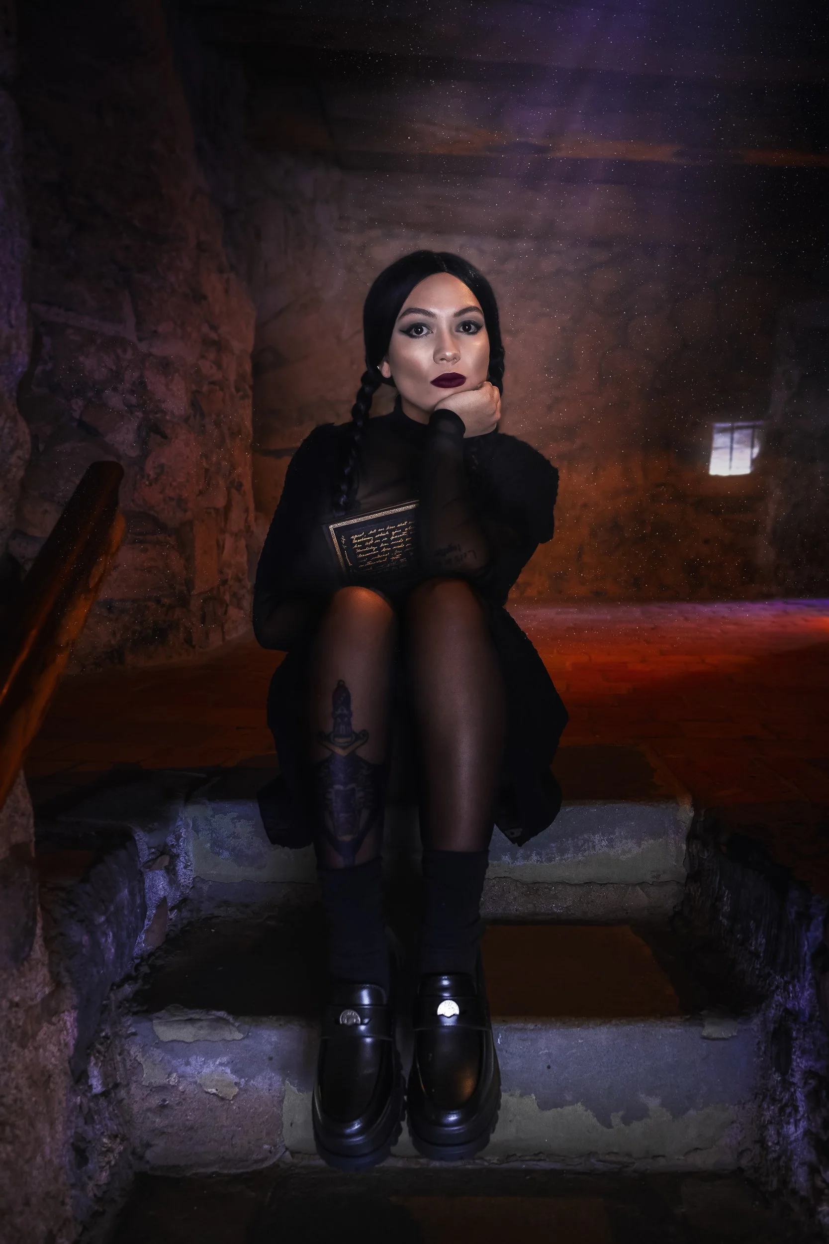 Une femme assise sur des escaliers en pierre dans une pièce sombre avec des murs en pierre, portant une robe noire, des collants noirs, des chaussures noires, avec ses cheveux noirs tressés et portant un maquillage sombre, tenant un livre dans ses ma