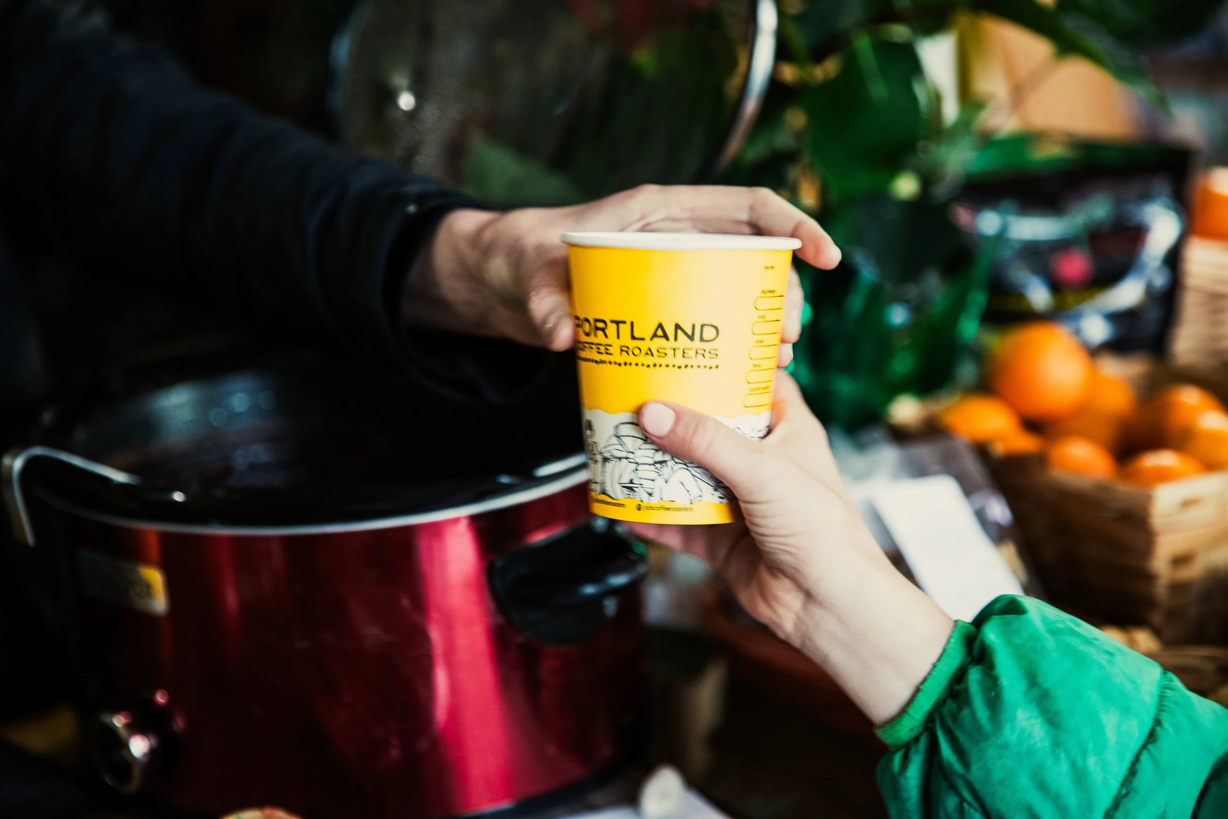 Person handing a yellow Portland Coffee Roasters cup to another person at a market or shop, with fresh produce and a red appliance in the background.