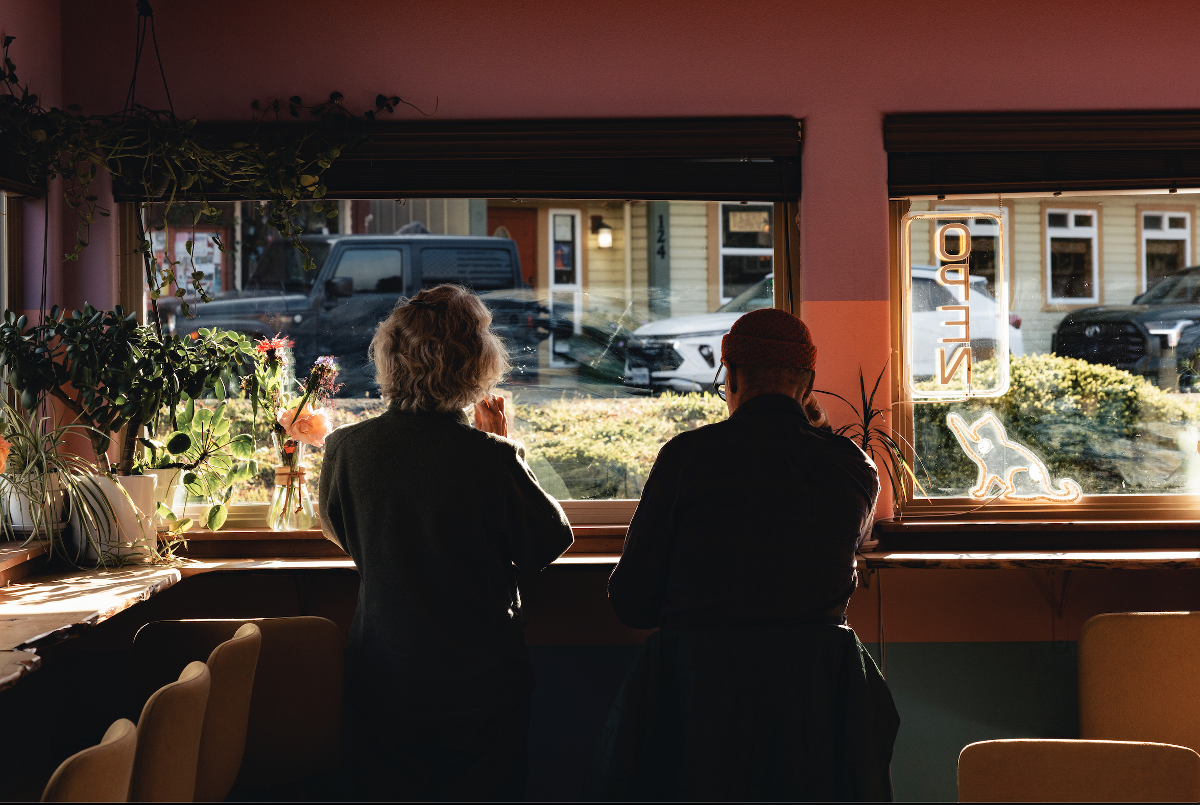Two people sitting at a counter inside a cafe, looking out the window at parked cars and buildings outside during daylight.