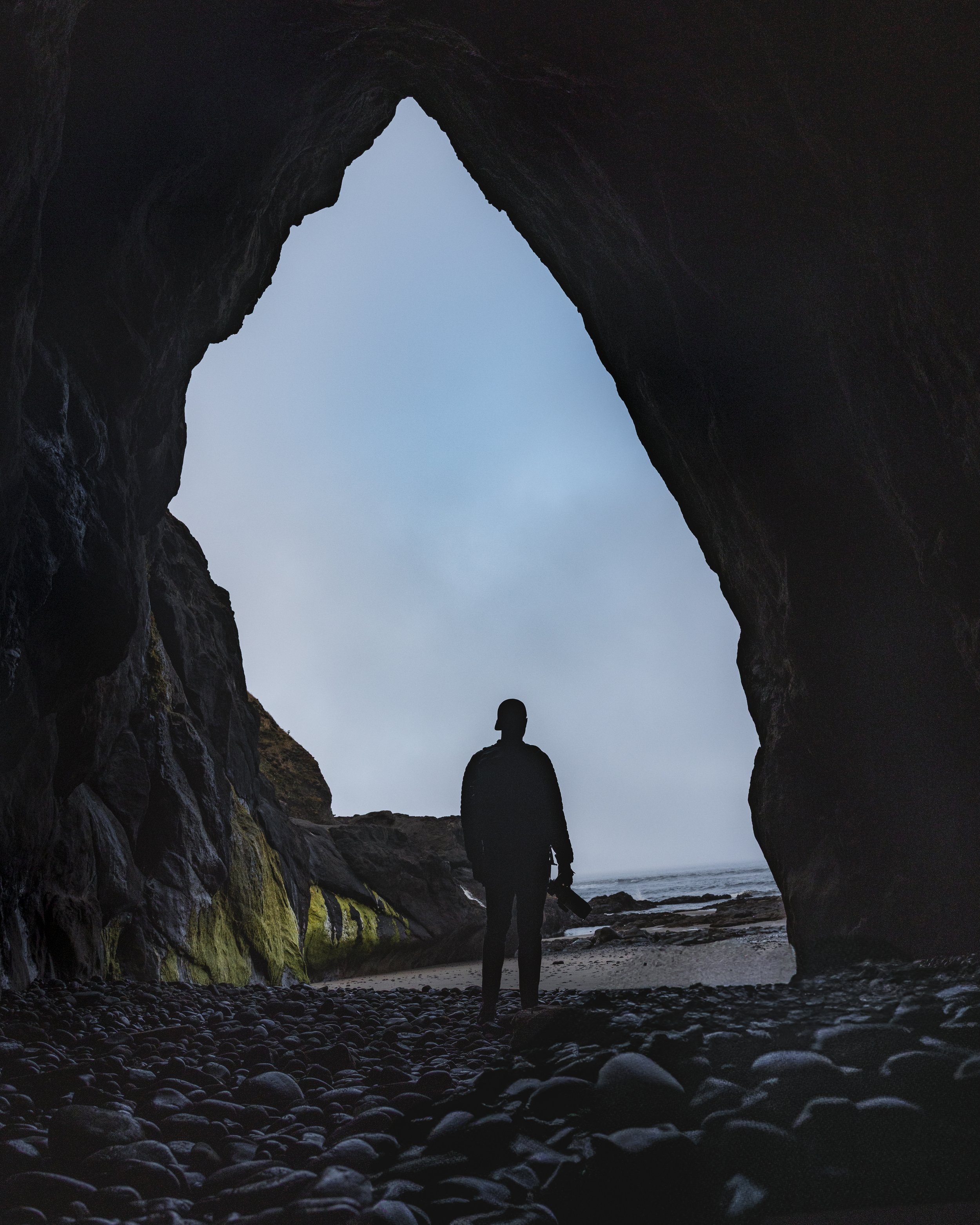Person standing at the entrance of a large sea cave, silhouette against the sky, with rocks and ocean visible outside.