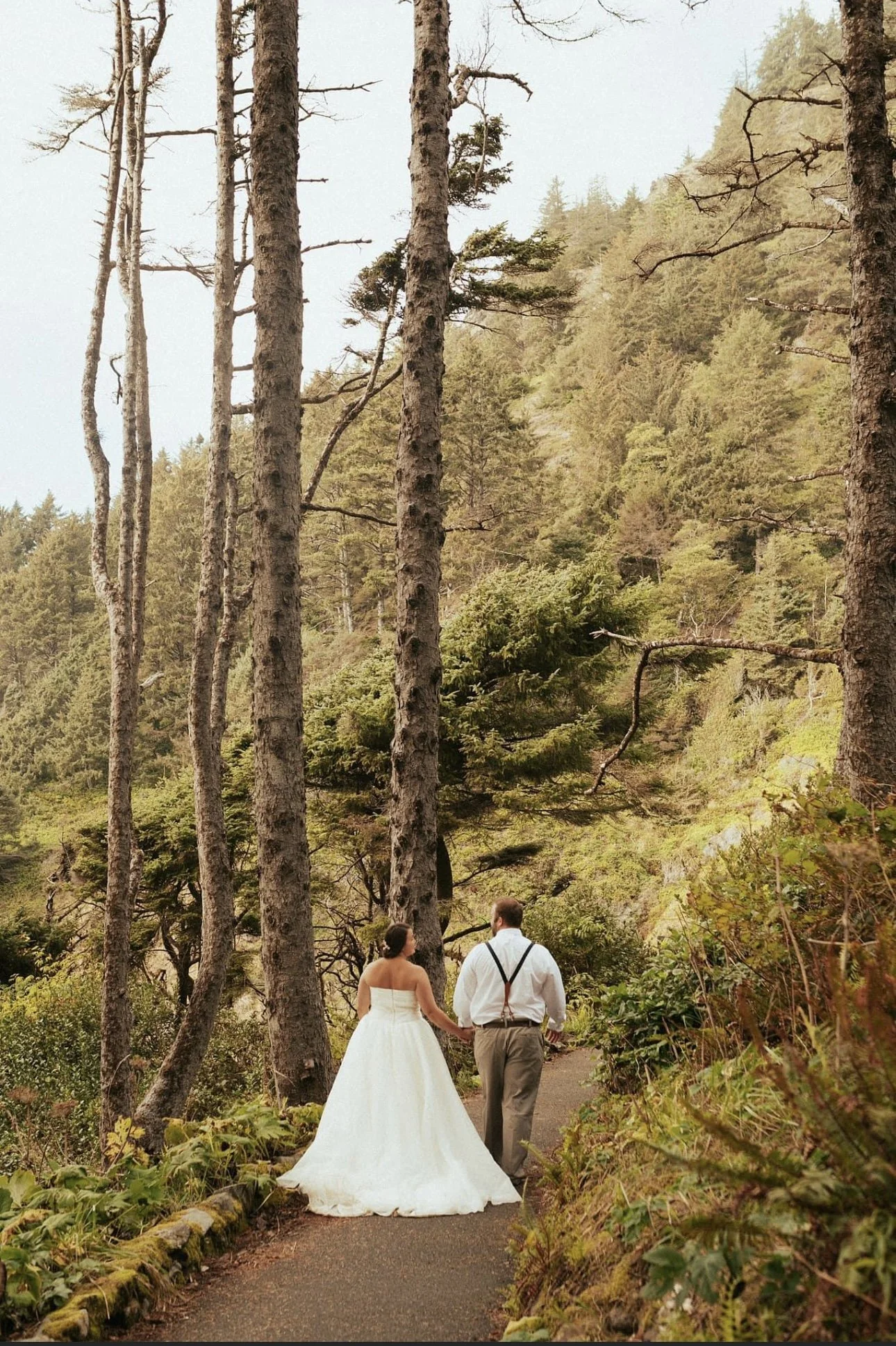A bride in a white wedding dress and a groom in a white shirt with suspenders walking hand in hand on a forest trail surrounded by tall trees and green foliage.