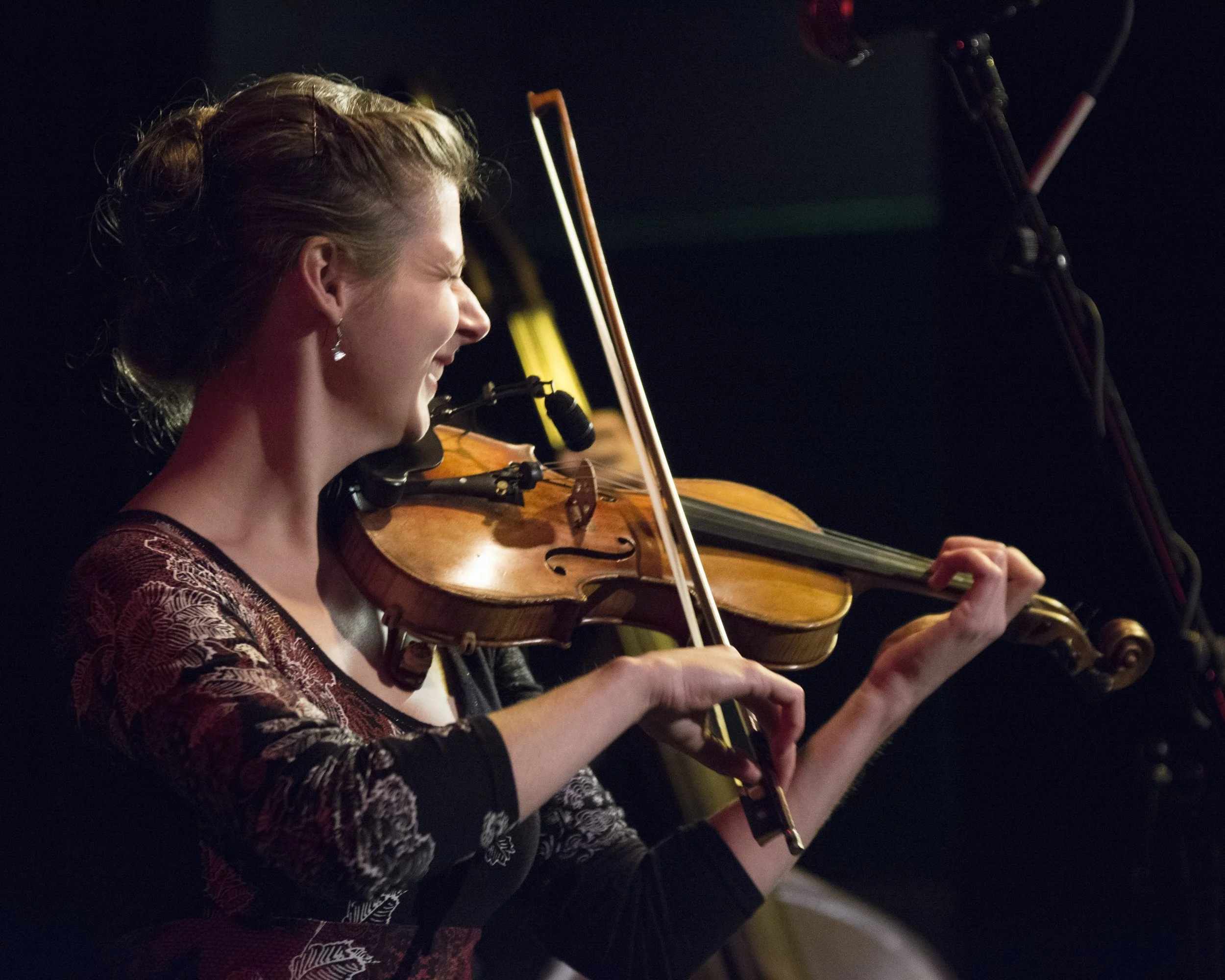 A woman playing the violin on stage, smiling with her eyes closed. She has her hair tied back and is wearing earrings and a patterned top. The background is dark and blurred.