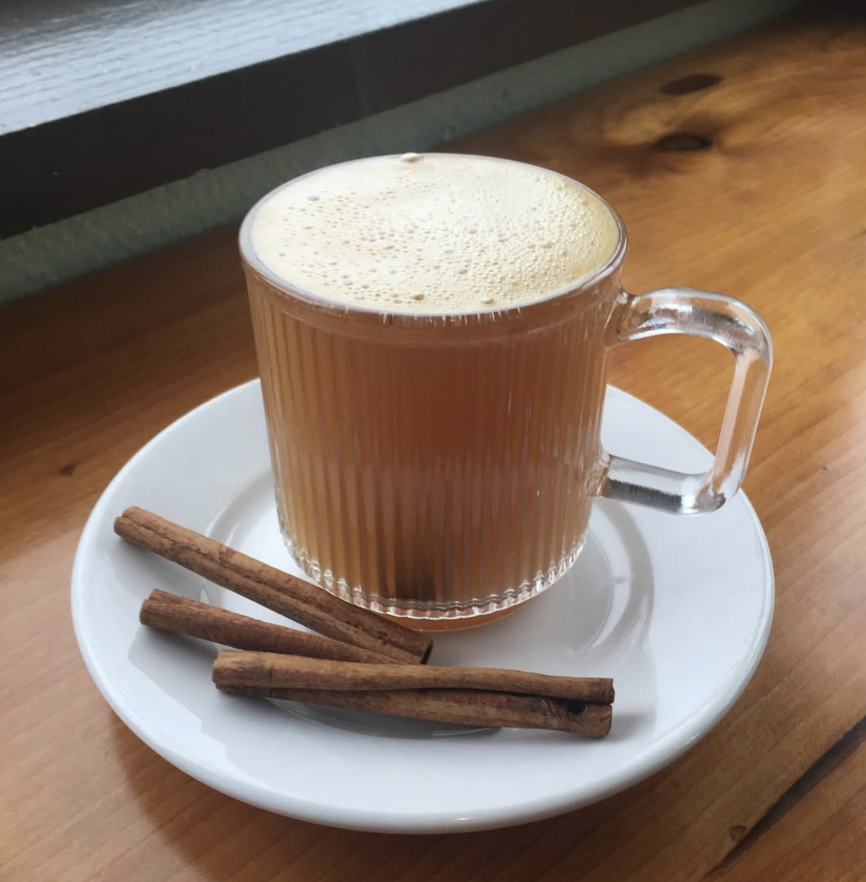 A glass mug of chai tea with frothy top on a white plate, garnished with three cinnamon sticks.