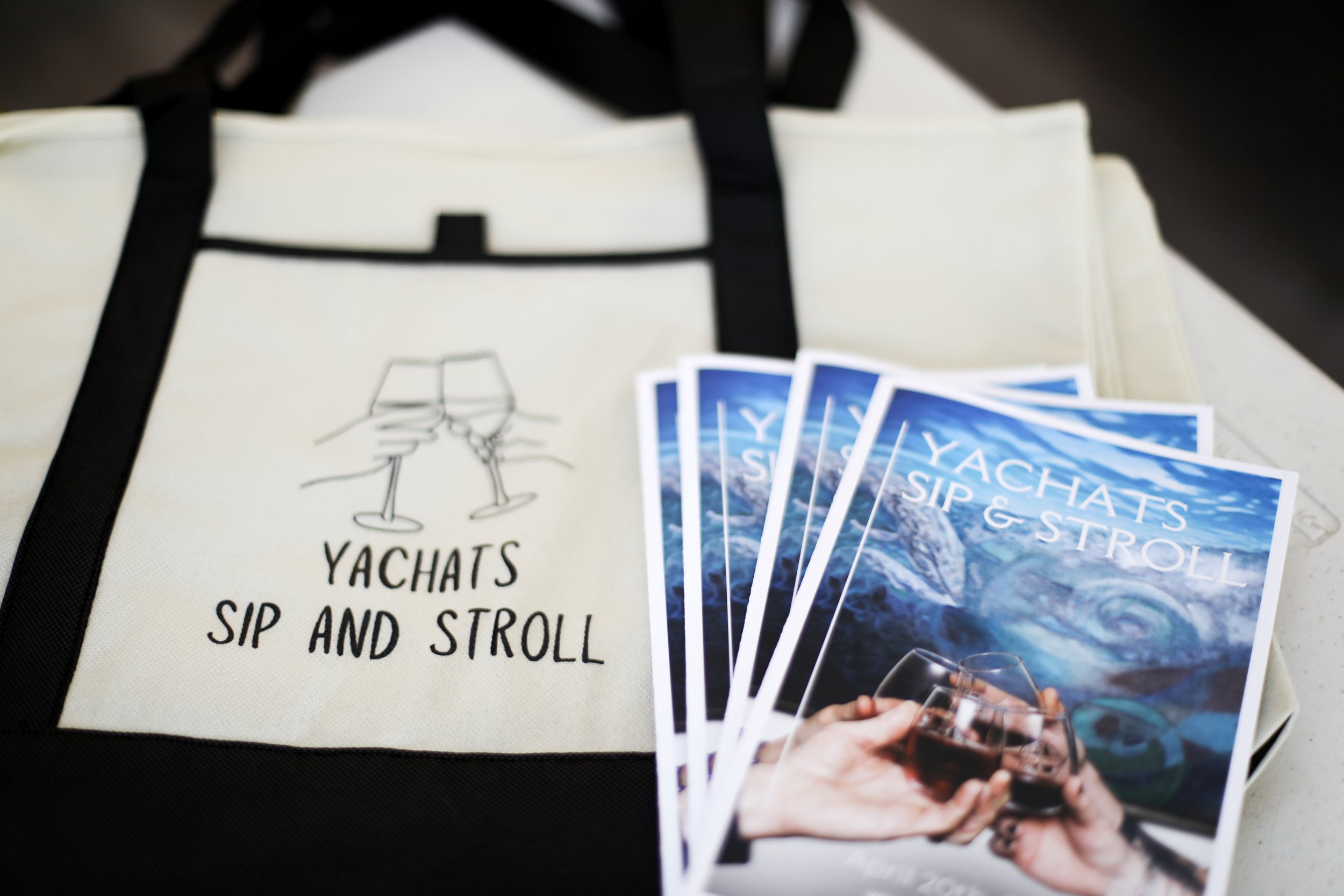 A tote bag with the logo 'Yachats Sip and Stroll' and several flyers for the same event, featuring a hand holding a glass of wine in front of a scenic ocean background.