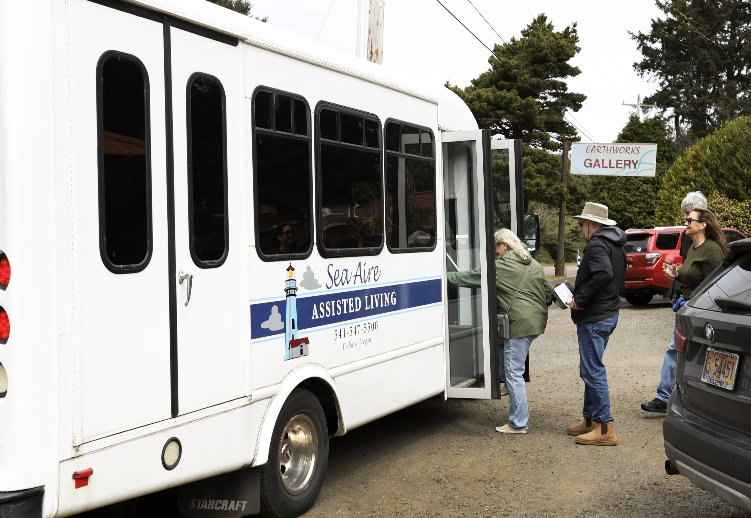 People lined up to board a white assisted living transportation vehicle labeled 'Sea Aire Assisted Living' with a lighthouse logo, outdoors near parked cars and trees, with a sign reading 'Earthworks Gallery' in the background.