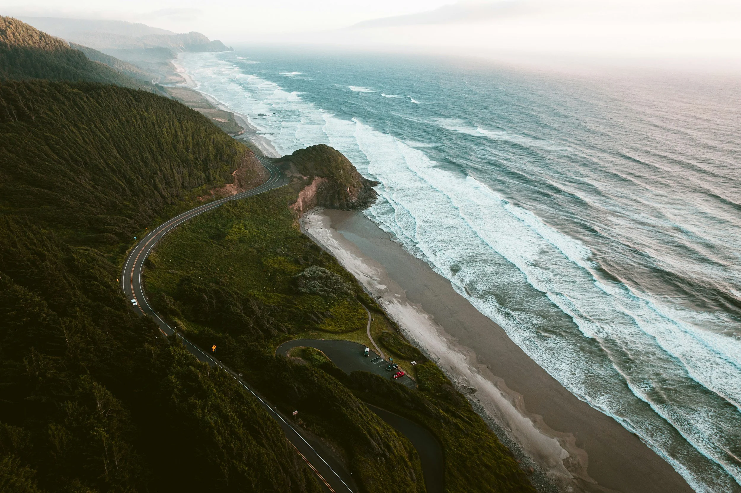 Aerial view of a winding coastal road along a forested hillside next to an ocean with waves breaking on the sandy beach.