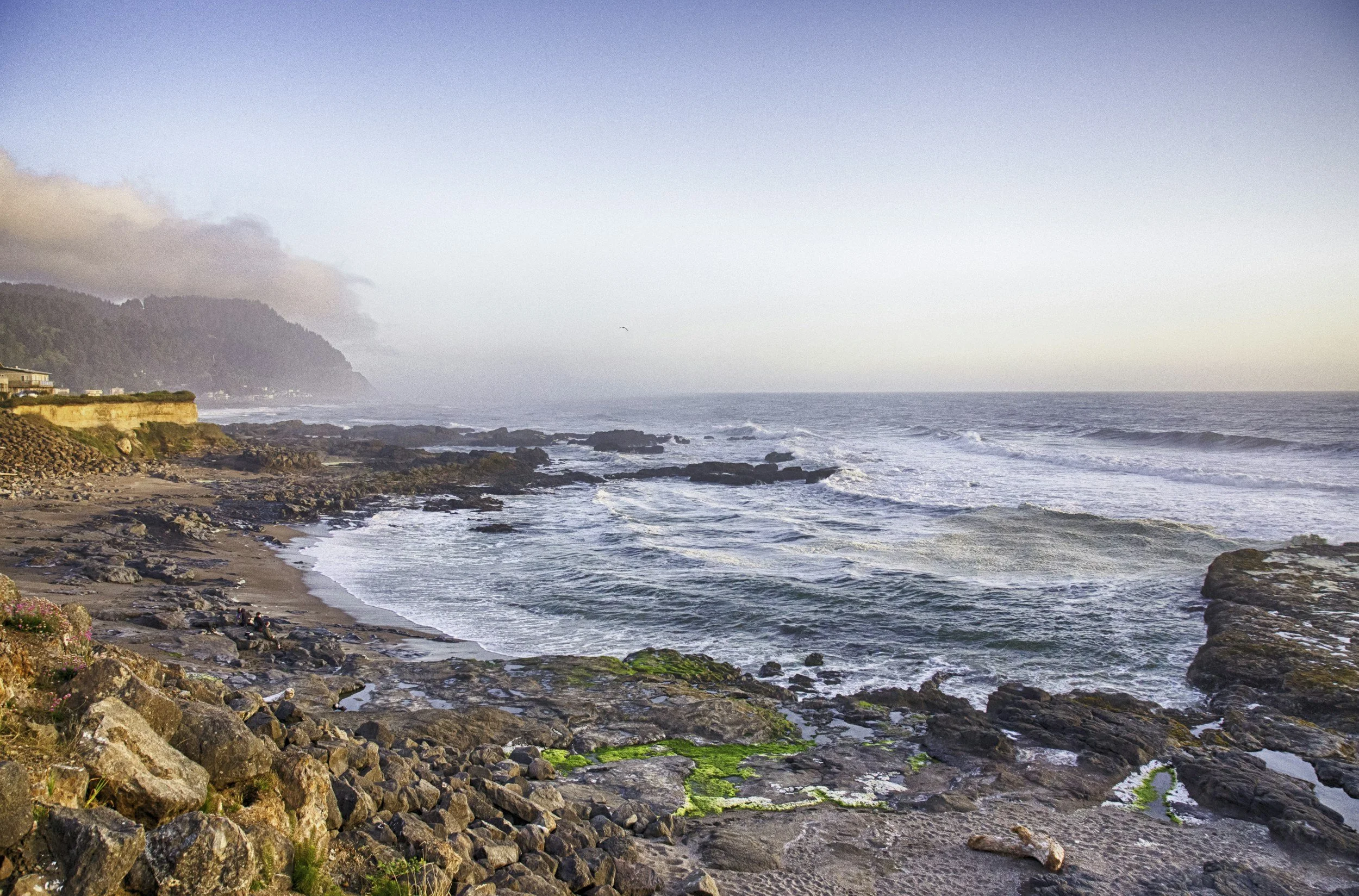 Scenic view of rocky beach with waves crashing, hillside houses, and foggy mountains in the distance during sunset.