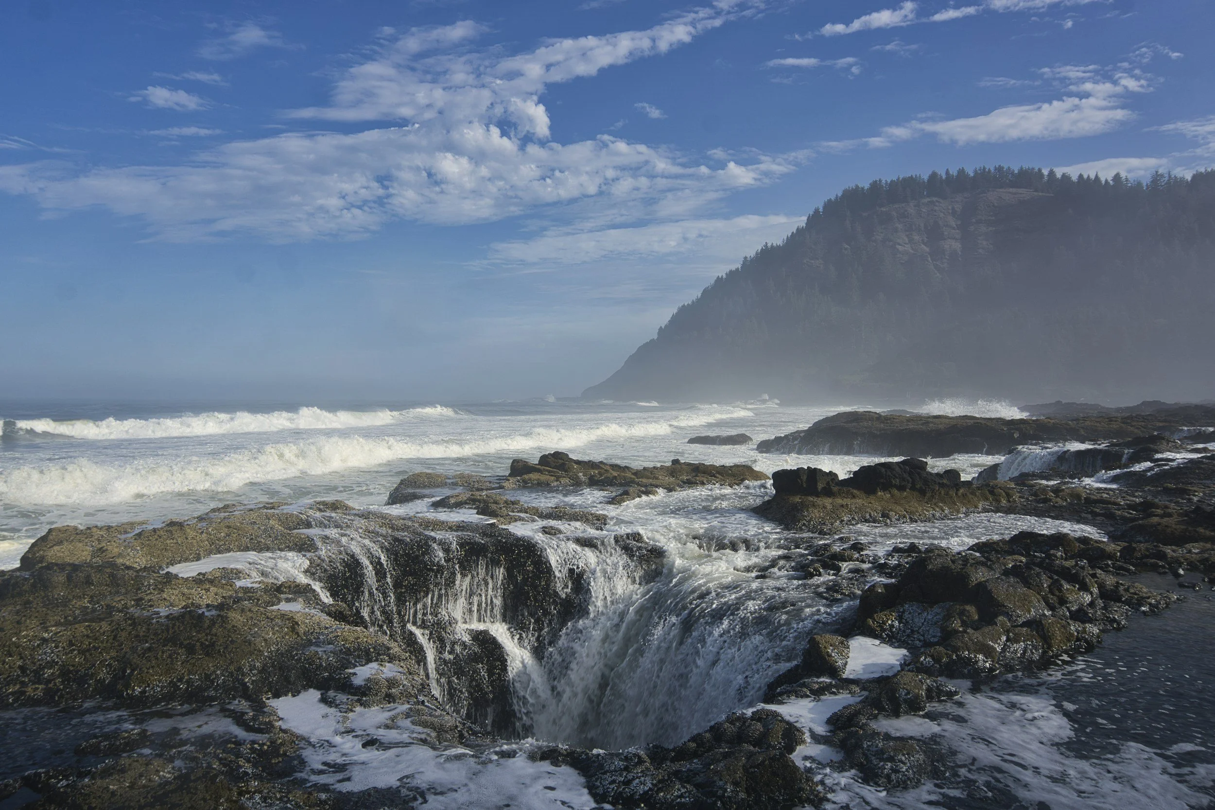Ocean waves crashing against rocky shoreline with a mountain and forest in the background, under a partly cloudy blue sky.