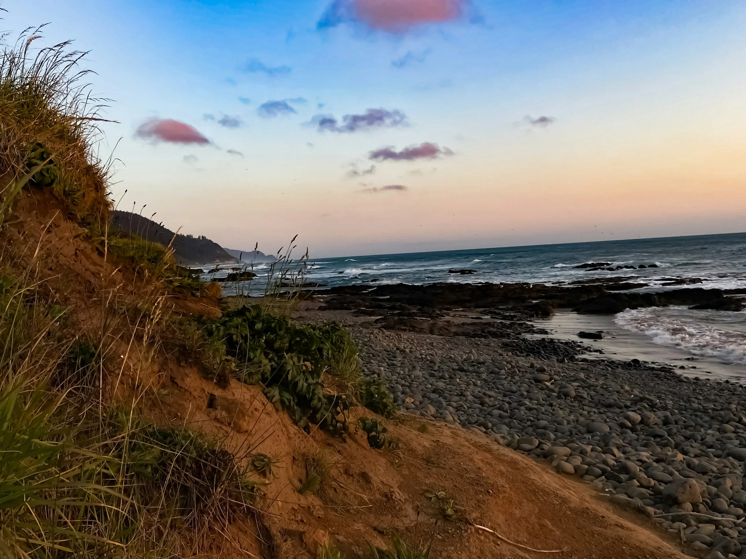 Sunset view of a rocky beach with waves, a distant shoreline, grassy sand dunes, and a partly cloudy sky.