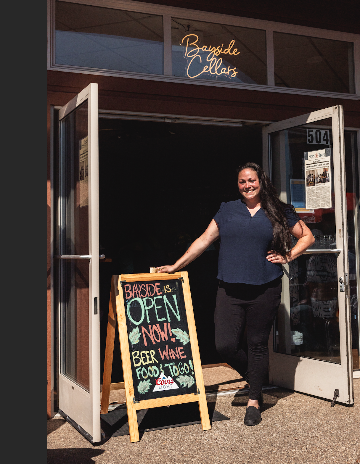 A woman standing at the entrance of Bayside Cellars, smiling. She is dressed in a dark blue top and black pants, with long dark hair. A chalkboard sign in front indicates the business is open and offers beer, wine, food, and to-go options.