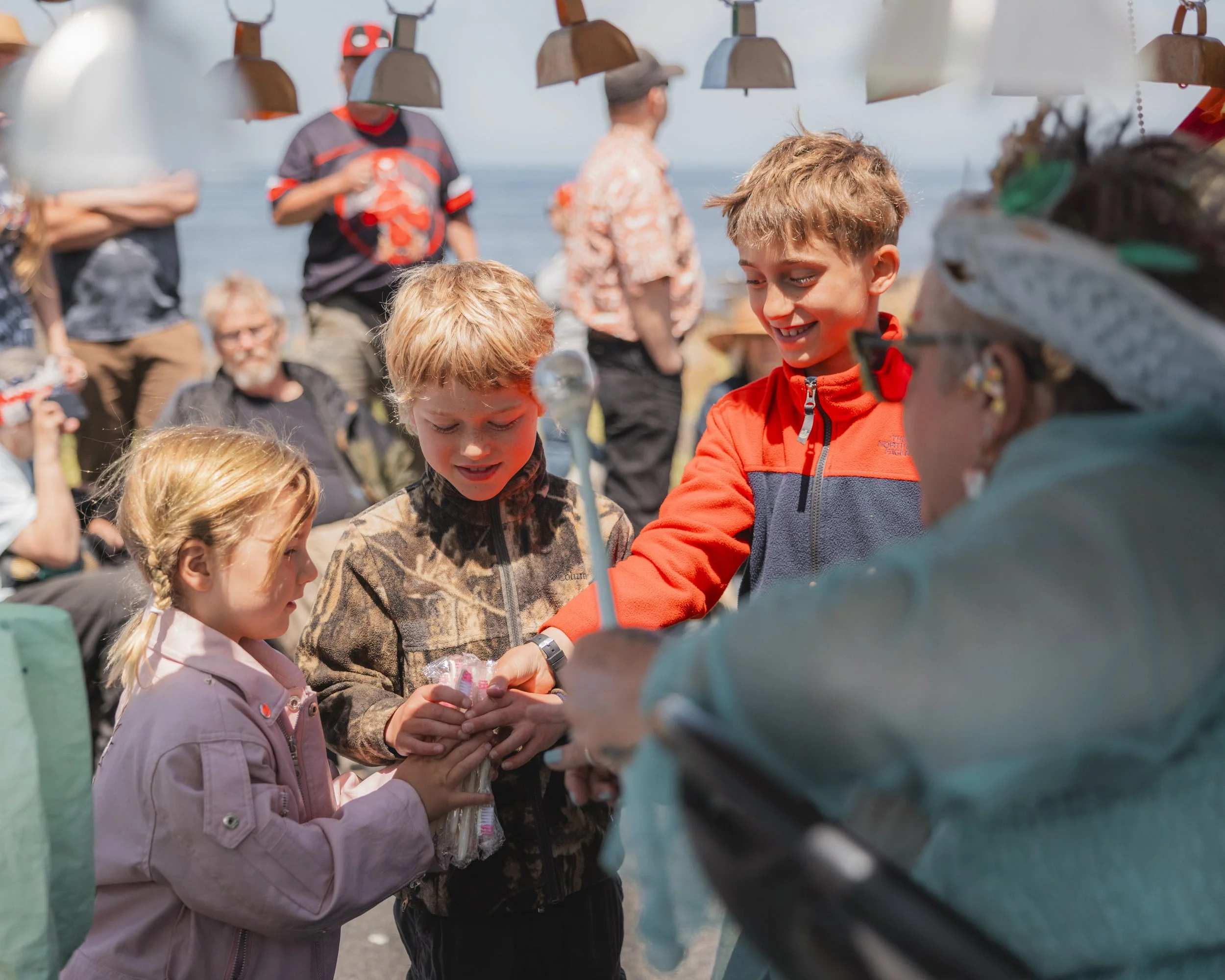 Children and adults gathered outdoors under hanging lamps, with the ocean in the background, as children receive or exchange items from an older woman wearing a hat with colorful decorations.