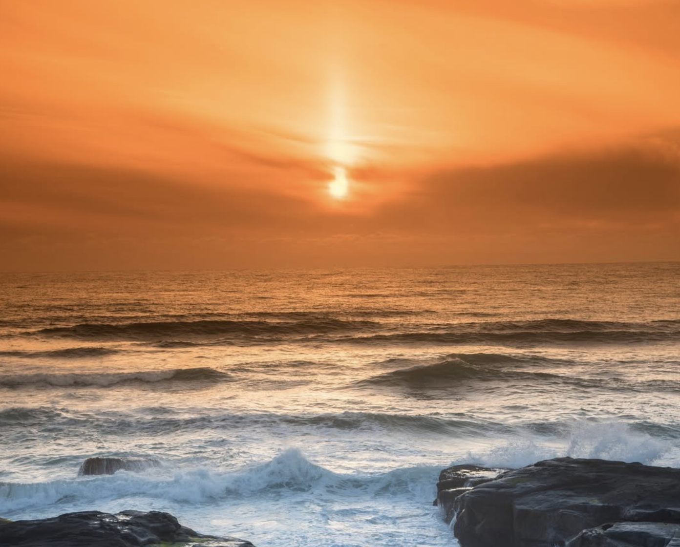 Sunset over the ocean with orange sky, waves crashing against rocks in the foreground.