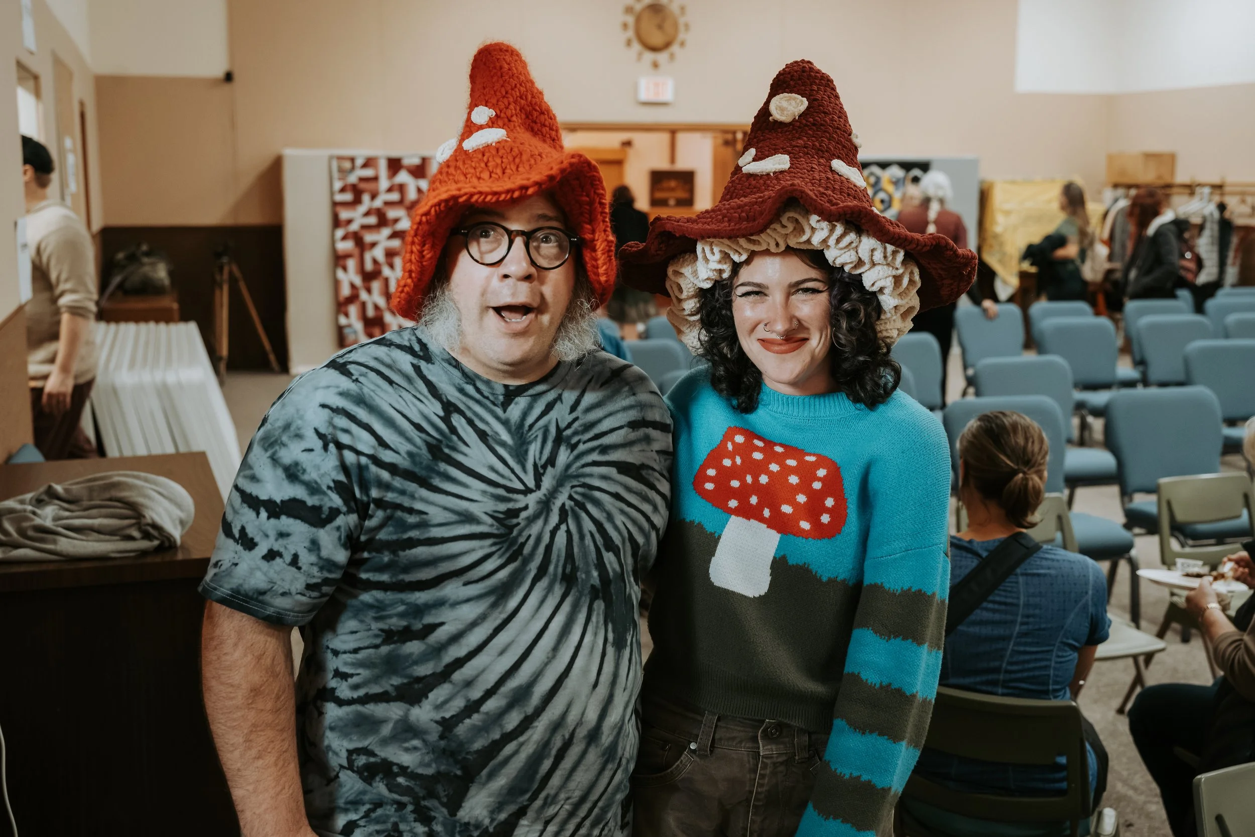 Two people wearing mushroom-shaped hats and colorful sweaters, standing in a room with chairs and other people in the background.