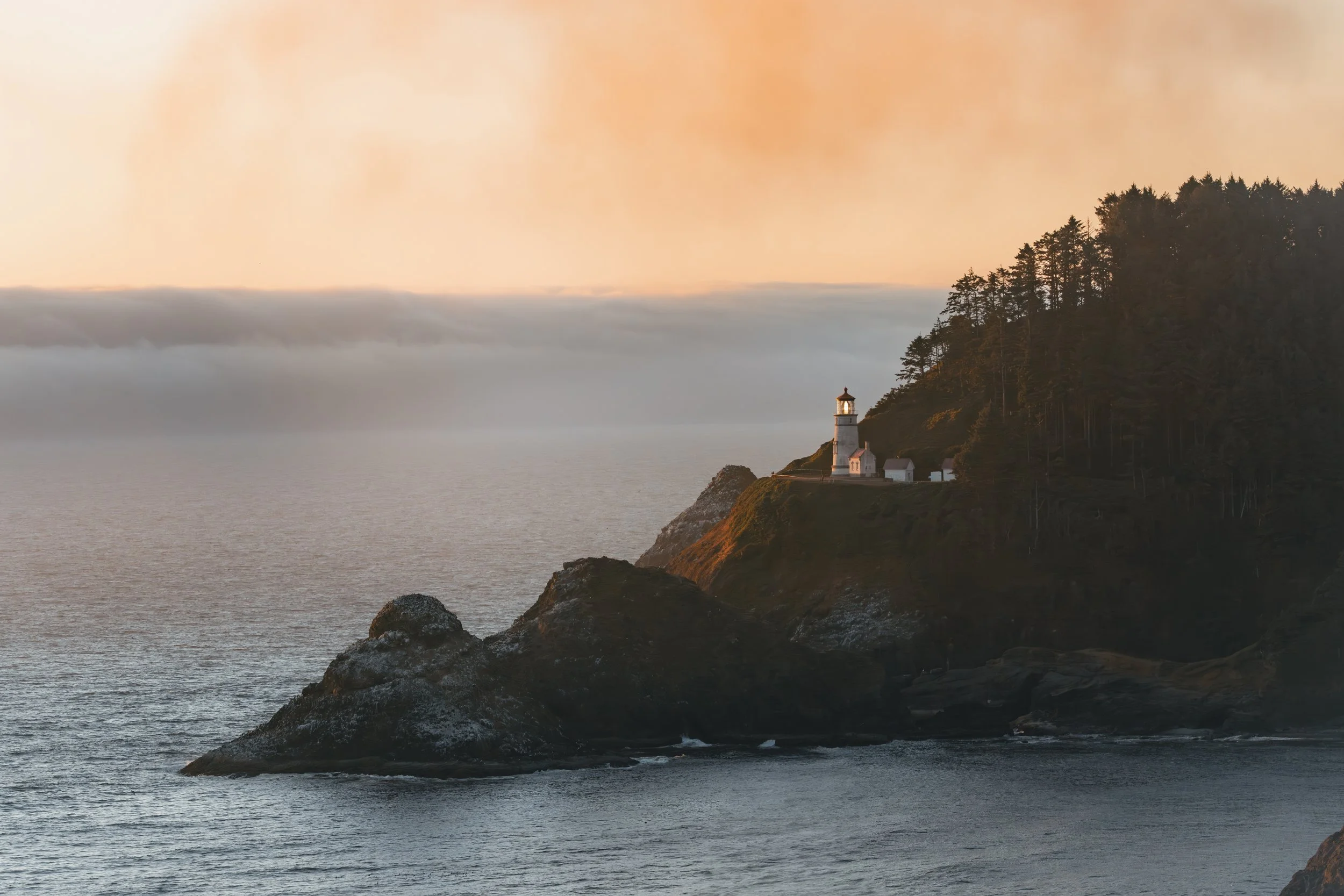 Sunset over water with lighthouse on a rocky cliff, surrounded by trees.