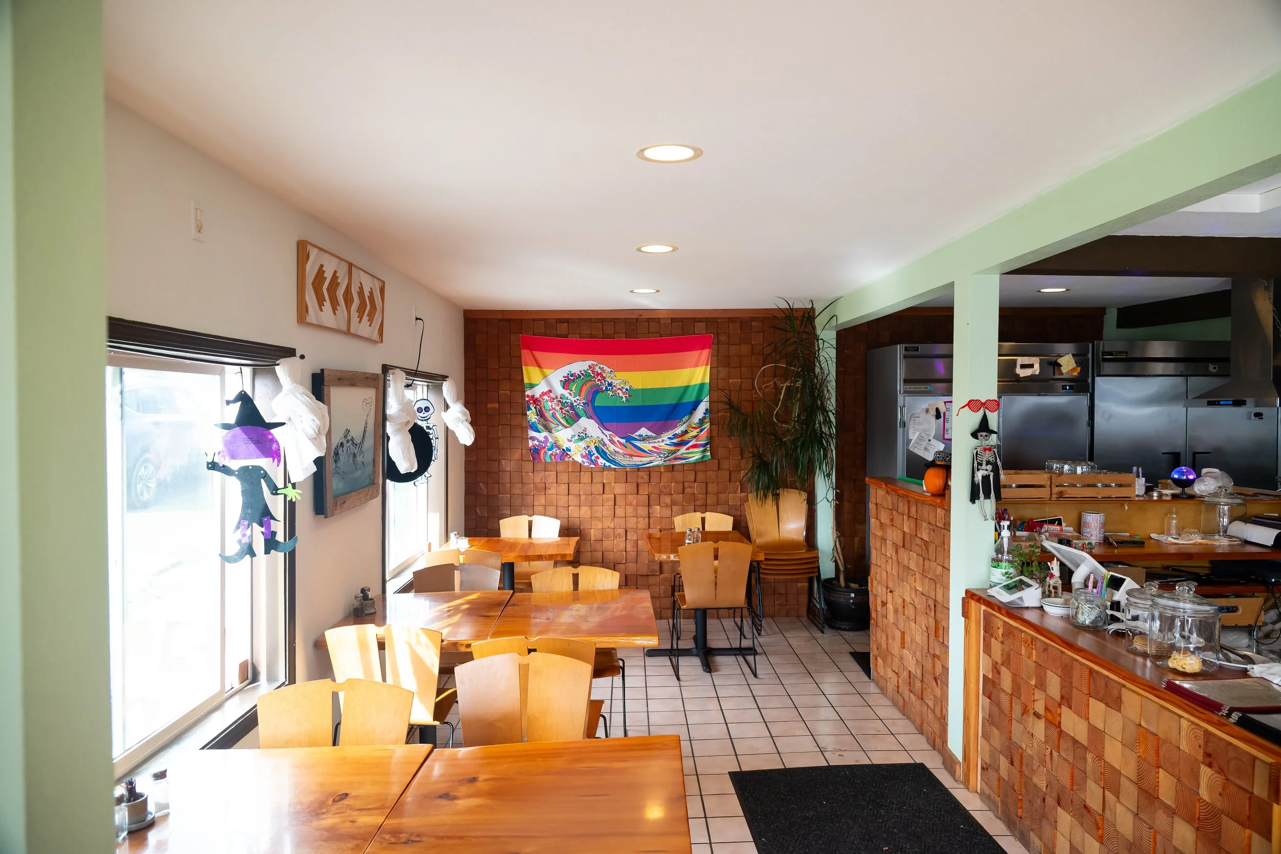 Interior of a cozy cafe with wooden tables, chairs, Halloween decorations, artwork, and a rainbow-colored wave tapestry on a brick wall, with visible kitchen area on the right.