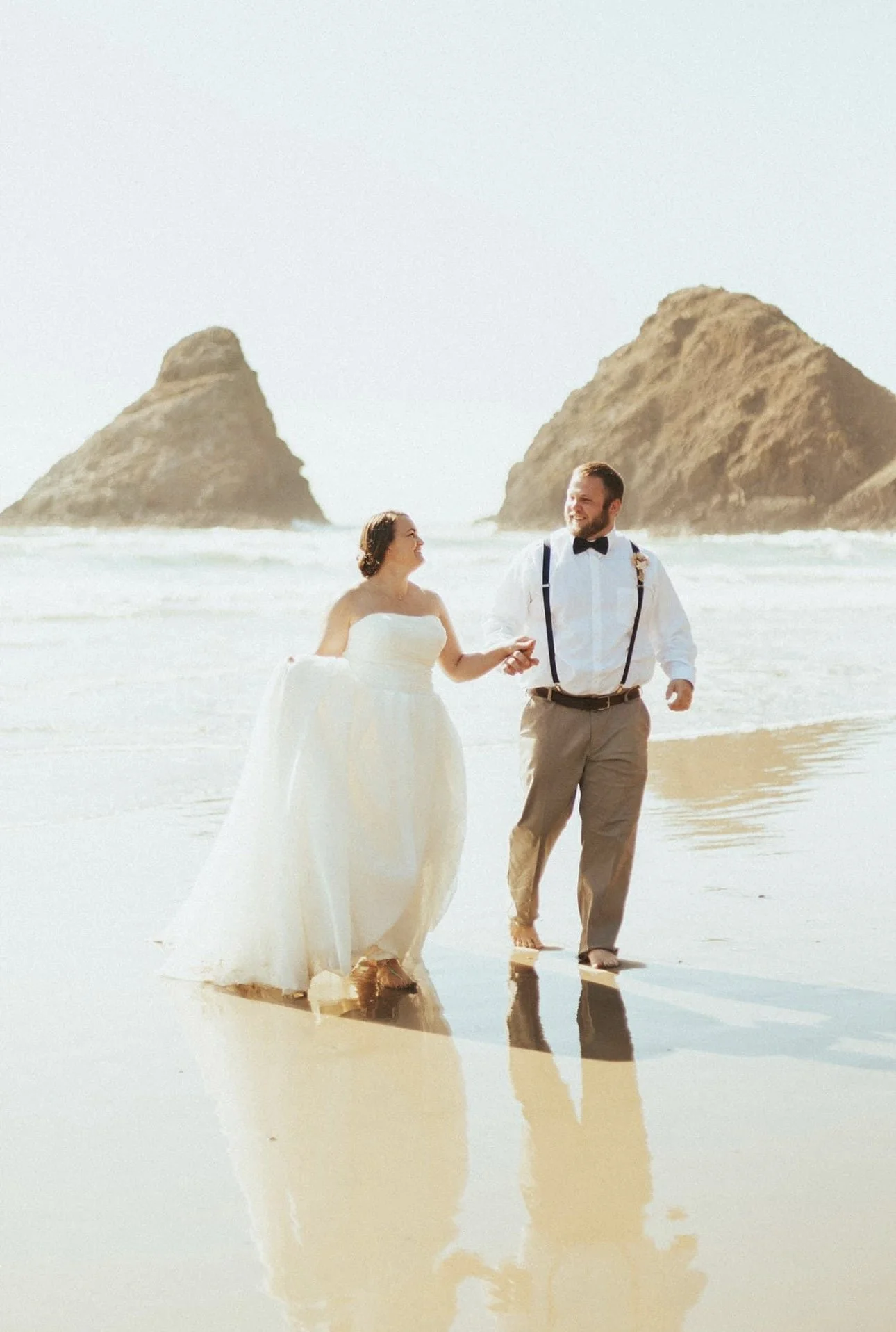 A newlywed couple walking along the beach at sunset, with large rocks in the ocean background.