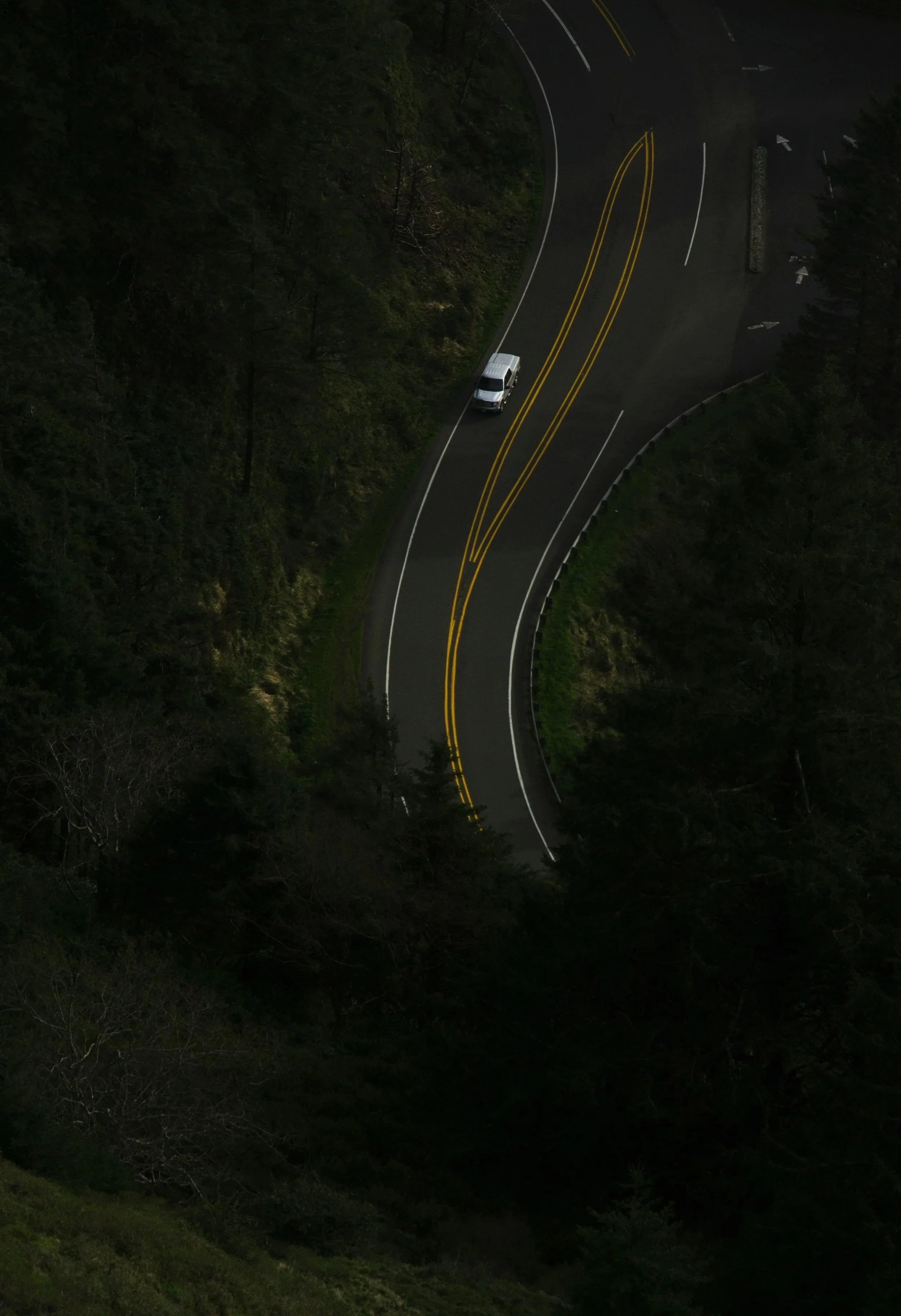 An aerial view of a winding mountain road with a white vehicle and trees on both sides.