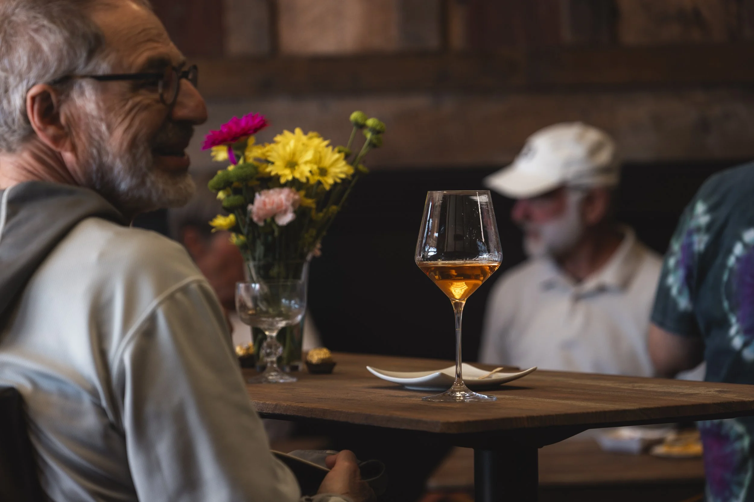 Man with glasses smiling, sitting at a table with a wine glass filled with rosé, a vase with pink, yellow, and green flowers, and small chocolates, in a dimly lit restaurant with wooden walls.
