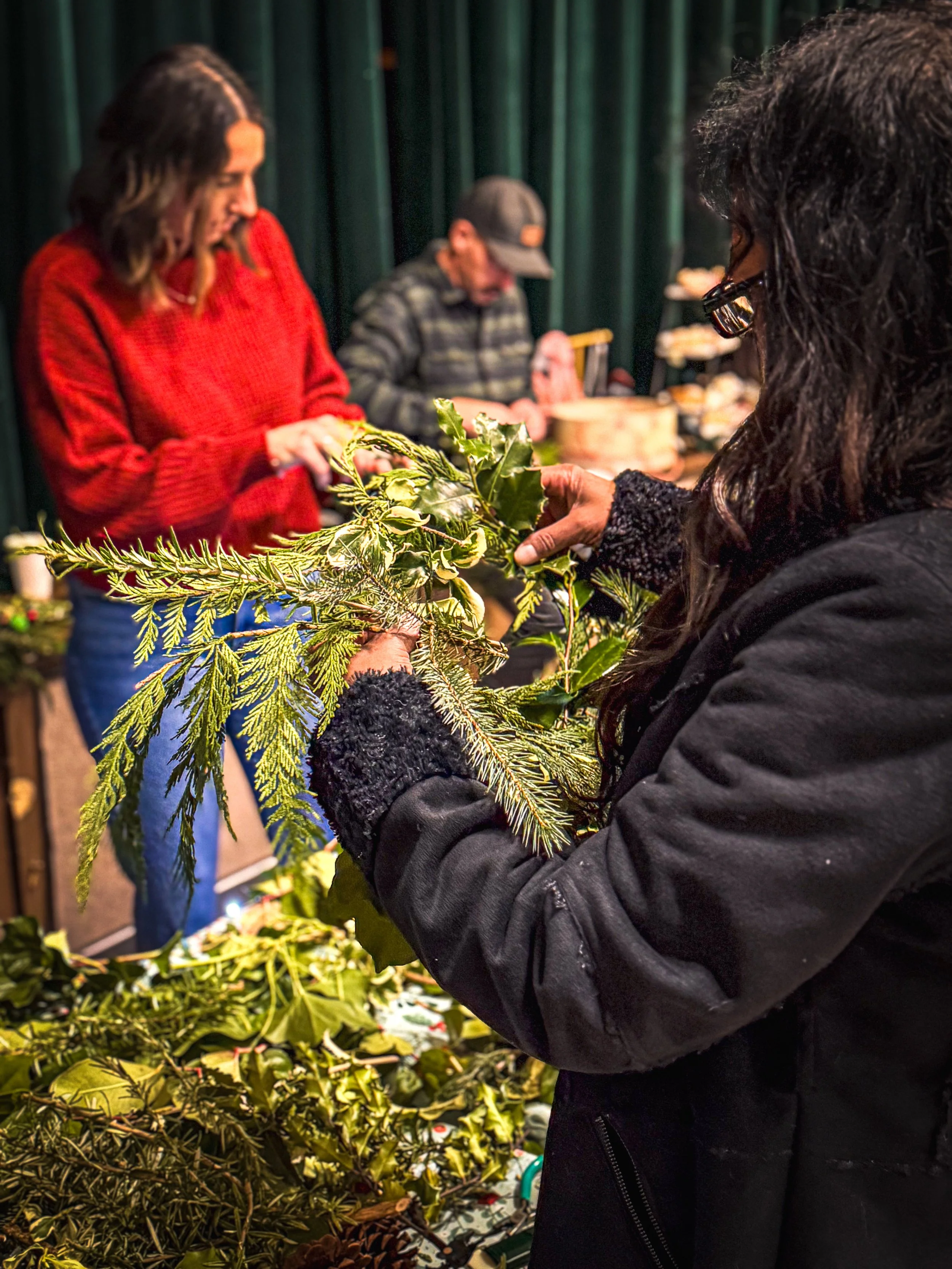 A woman wearing glasses and a black coat is holding and examining green holiday foliage, possibly preparing for a decoration. In the background, two other people are looking at similar items, with a table laden with more greenery and supplies.