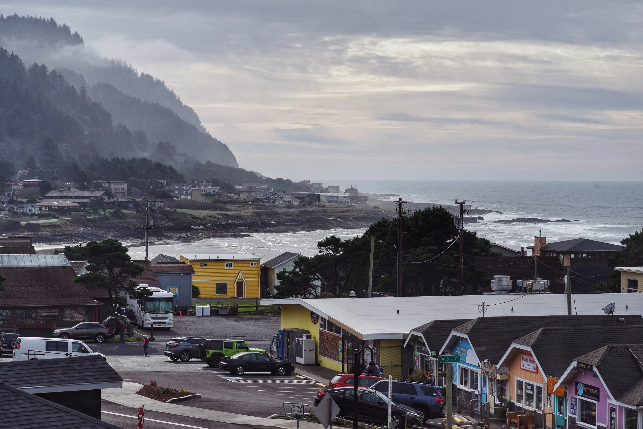 Coastal town with colorful shops, parked cars, and a cloudy sky over the ocean, with houses on a hillside in the background.