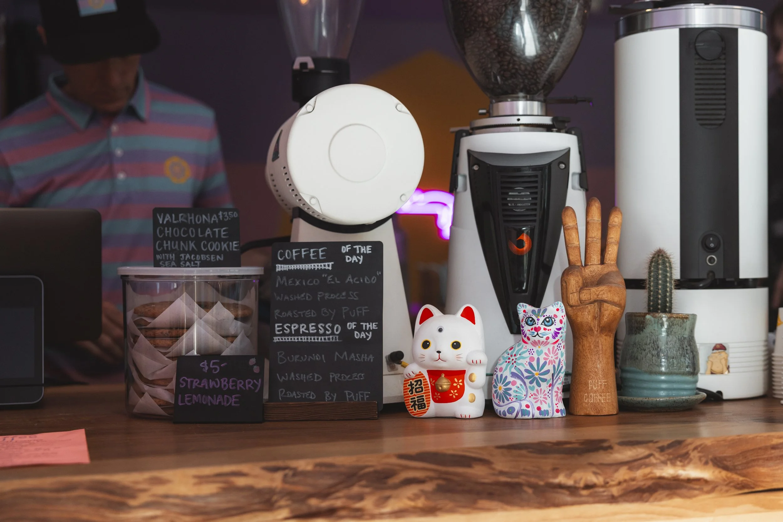 Coffee shop counter with various items: a white coffee grinder, a brown wooden peace hand sign, two decorative cat figurines, a cactus in a ceramic pot, and a white beverage dispenser. Handwritten signs display prices and coffee of the day. A person 