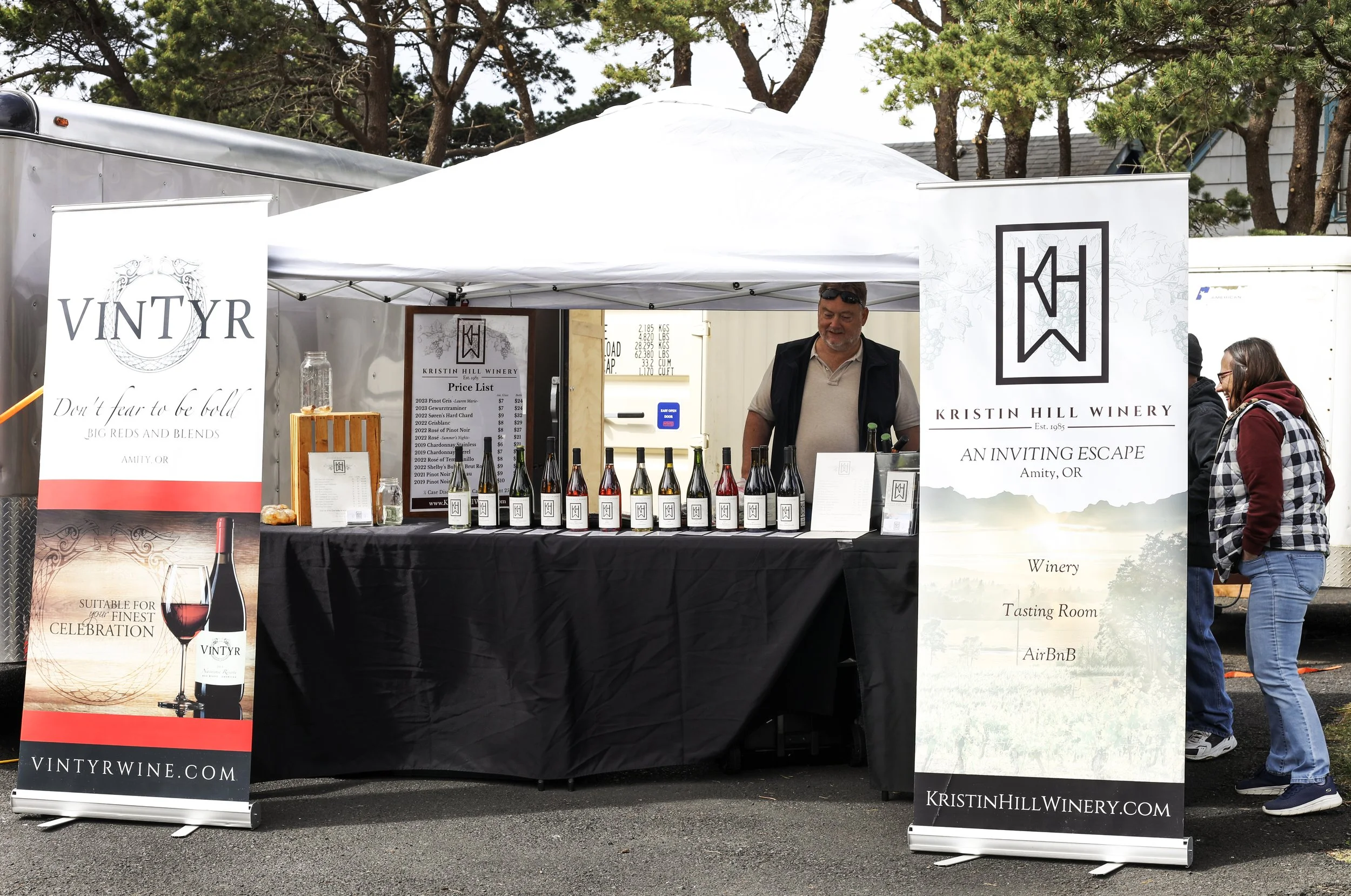 At an outdoor wine tasting booth for Kristin Hill Winery in Amity, Oregon, a man is standing behind a table displaying bottles of wine with a woman and a few others browsing in the background.