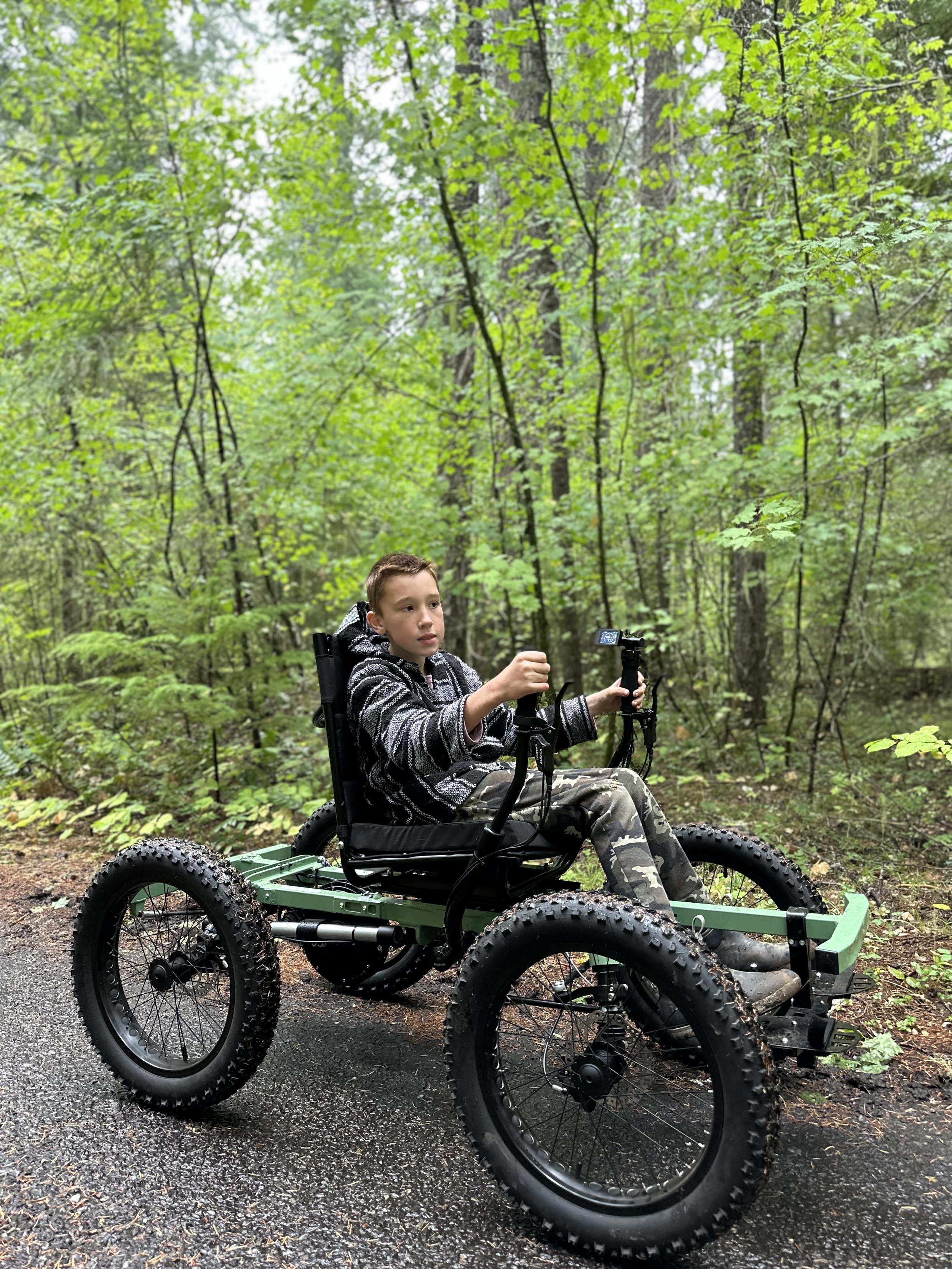 A young boy in a striped hoodie and camouflage pants sitting in a four-wheeled electric off-road wheelchair on a wet forest trail, surrounded by green trees and foliage.
