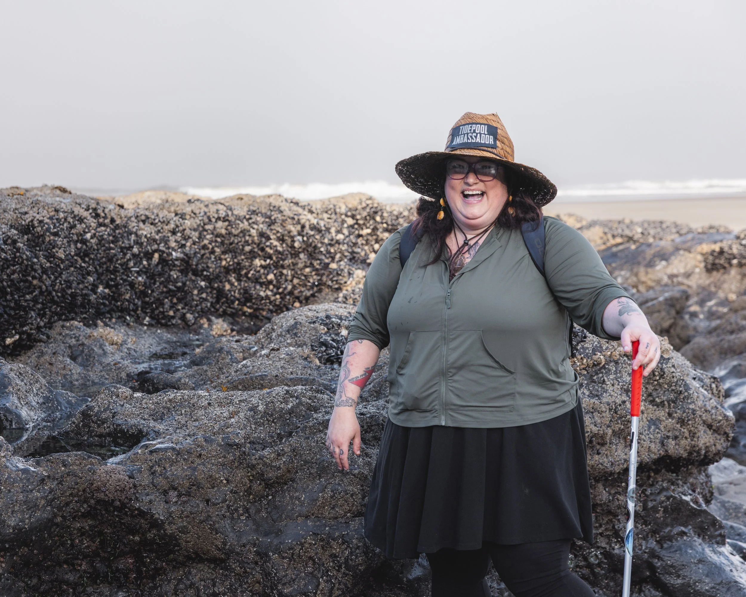 A woman standing on rocks at the beach, laughing, wearing a wide-brimmed hat, glasses, a green jacket, a black skirt, and holding a walking stick.